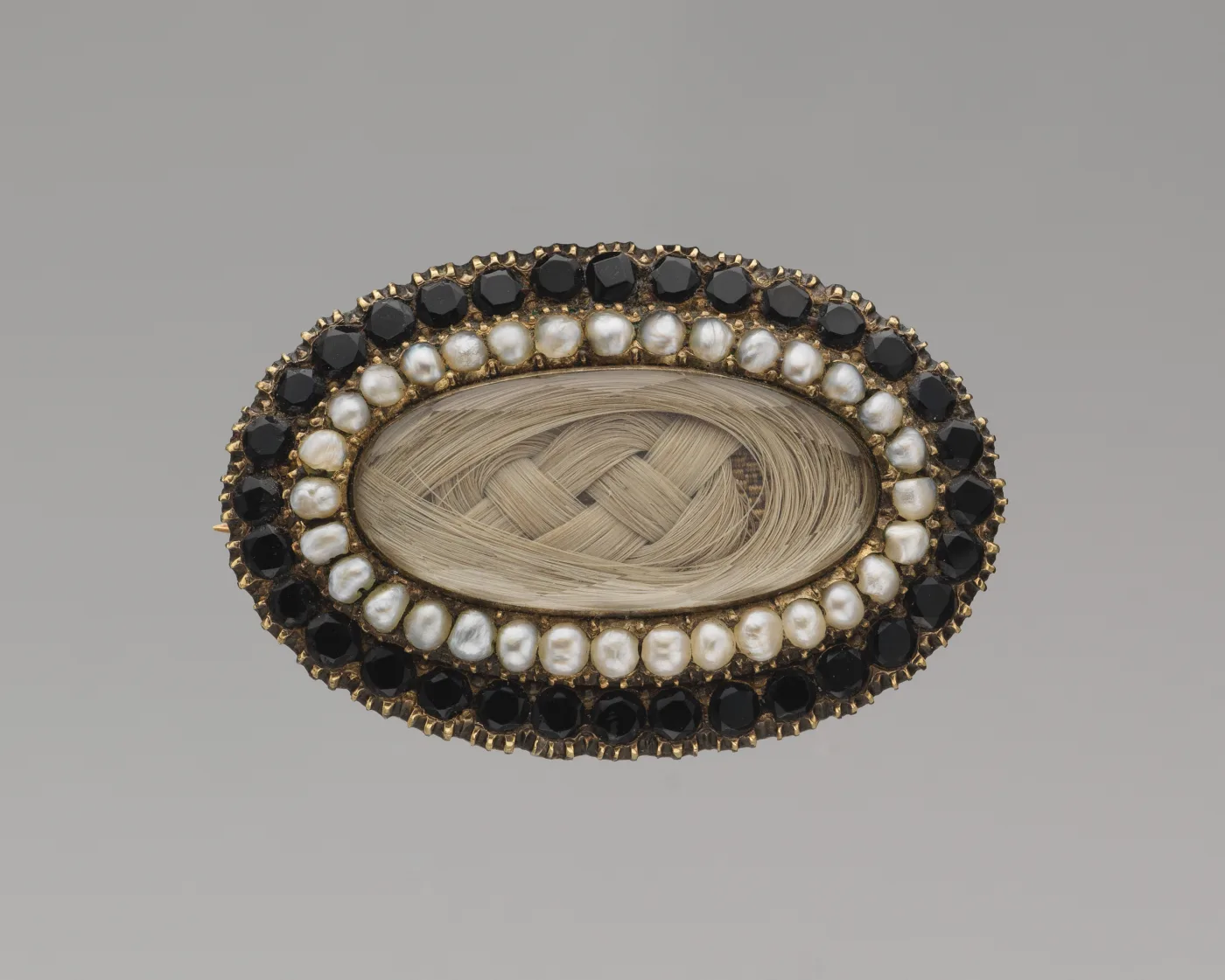 Oval gold brooch with a glazed centre panel of plaited blonde hair, ringed by a row of seed pearls and an outer row of faceted black stones