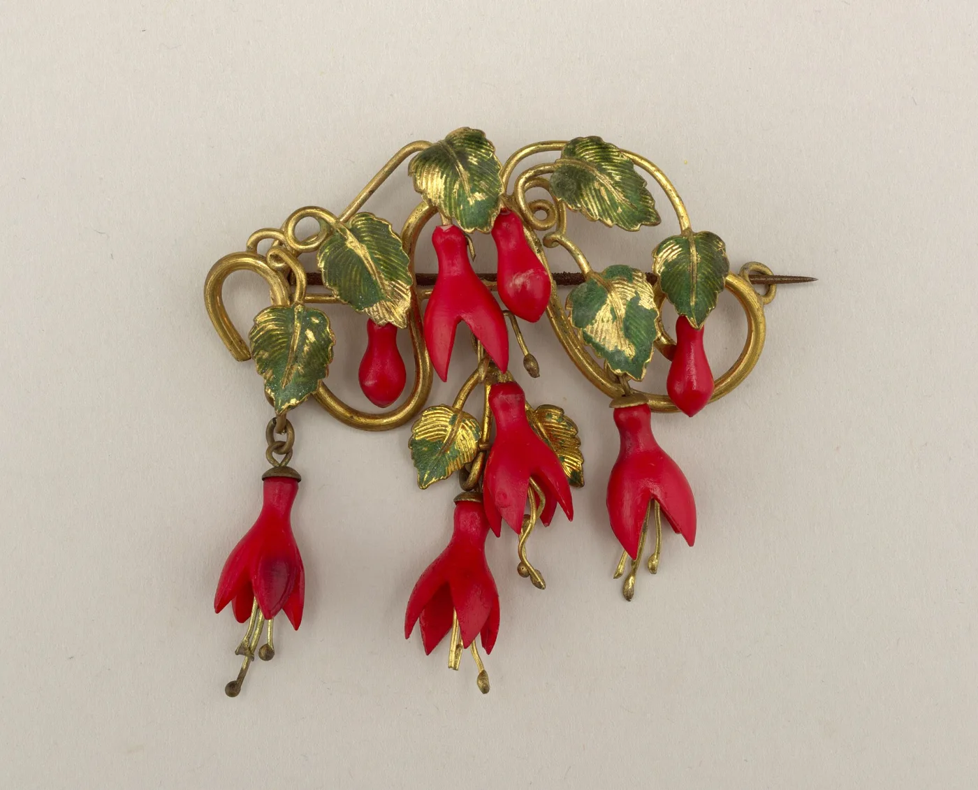 Gilt-metal brooch formed as a spray of fuchsia flowers with red carved blossoms and green-tinted leaves, the steel pin stem projecting to the right