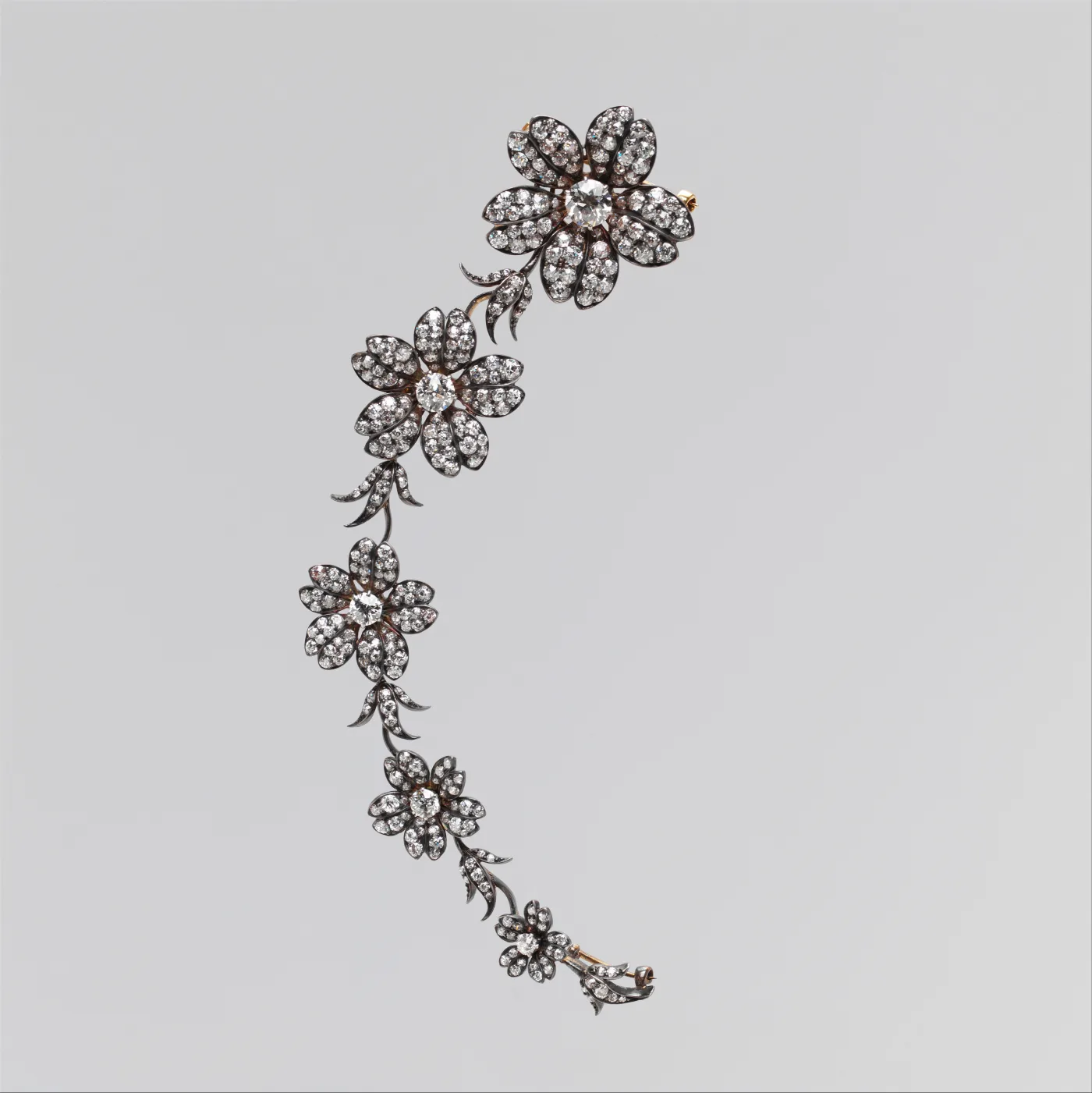 Curved spray of five diamond-set flowers with leaves, the diamonds mounted in dark silver collets backed by yellow gold, with pin fittings at each end