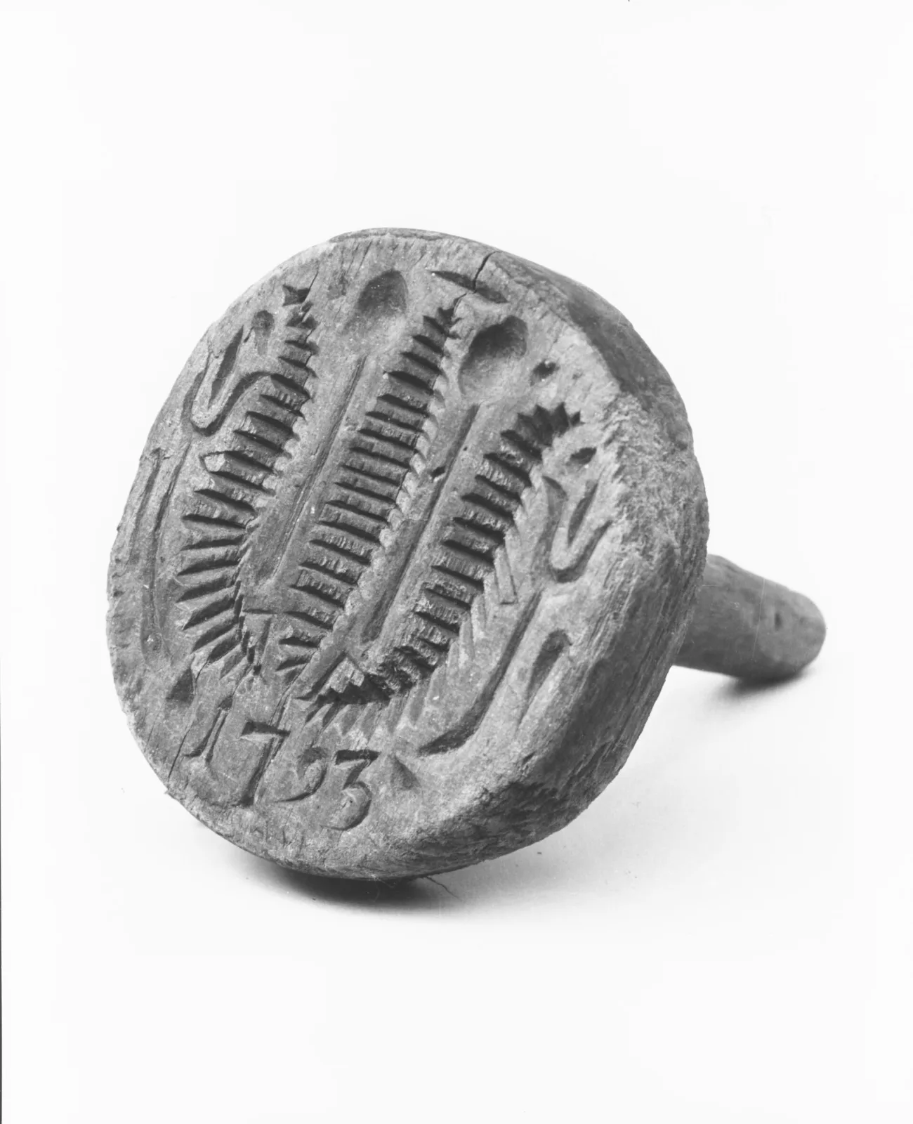 Round wooden butter stamp head with a short turned handle behind it, its face carved with ears of wheat and a date, photographed on white