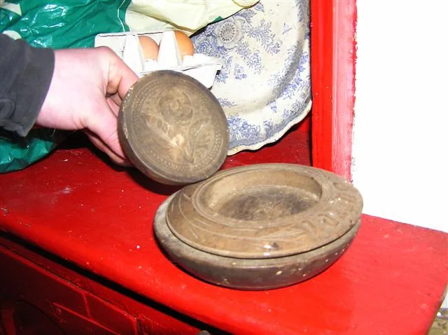 A pair of hands holding a round wooden butter stamp above a red-painted table, with a second wooden mold lying beside it