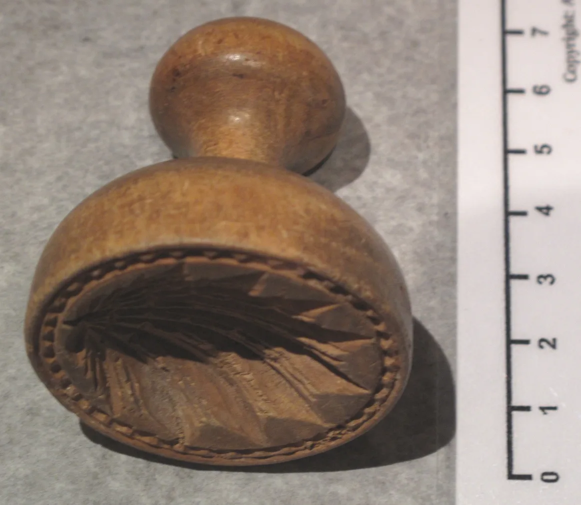 Round wooden butter stamp with a leaf design carved into its face, lying beside its short handle with a centimetre ruler along one edge