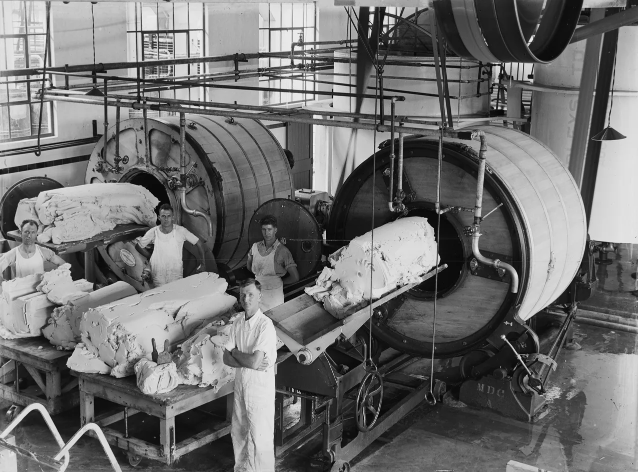 Black-and-white photograph of four workers in white overalls beside a row of large rotating wooden butter churns in a dairy factory, with blocks of butter on a bench