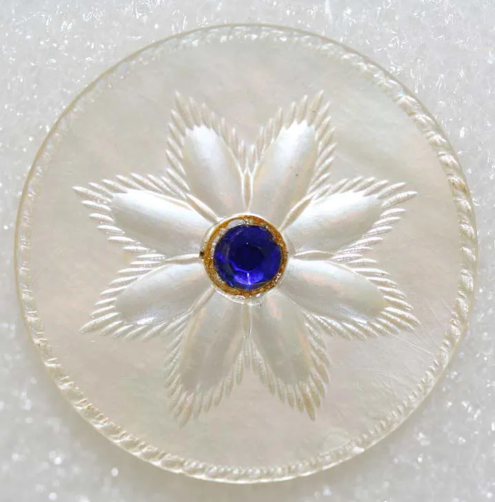 Mother-of-pearl button carved in relief as a six-petalled flower with engraved veining, a faceted blue stone in a gilt collet at its centre, inside an engraved rope border
