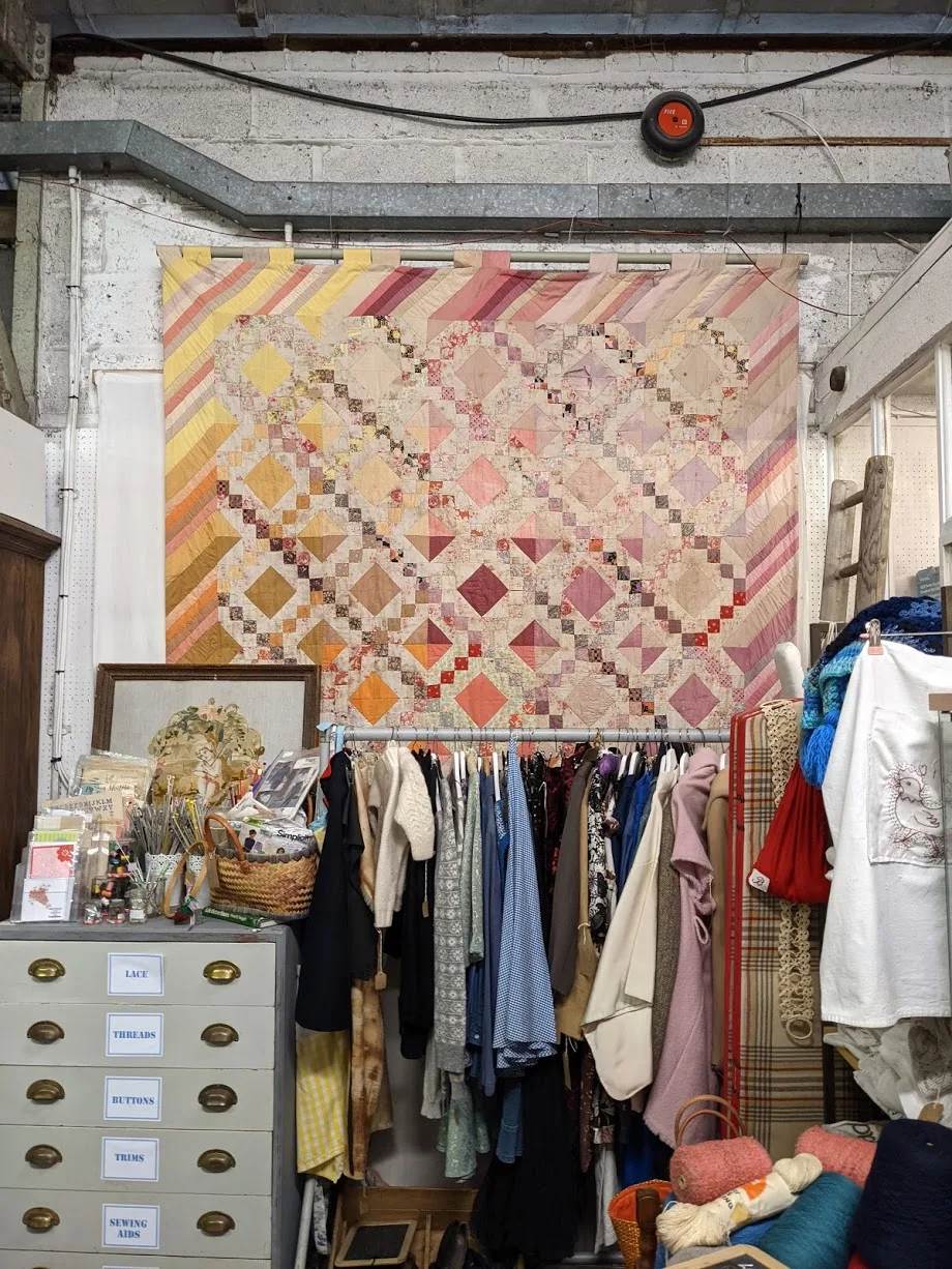 Worn patchwork quilt in graduated colours hanging as a backdrop on a market stall, with clothing and a bank of small drawers below it