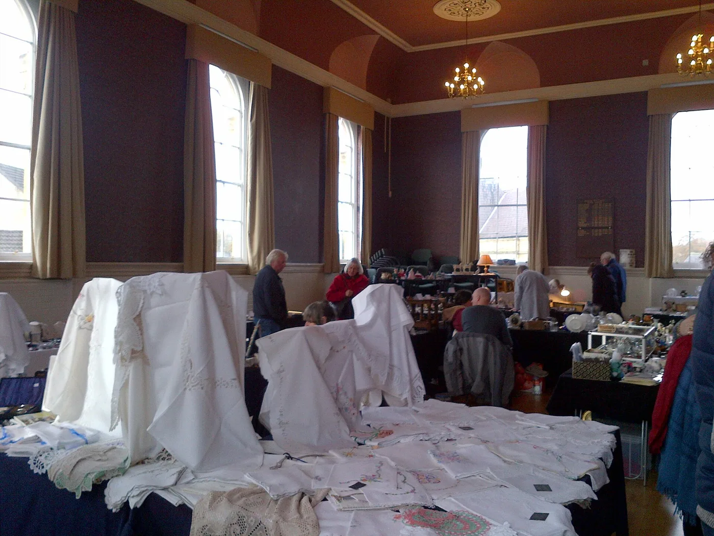 Small-town antiques fair in a plain municipal hall, with cloth-covered tables of stock and a handful of visitors