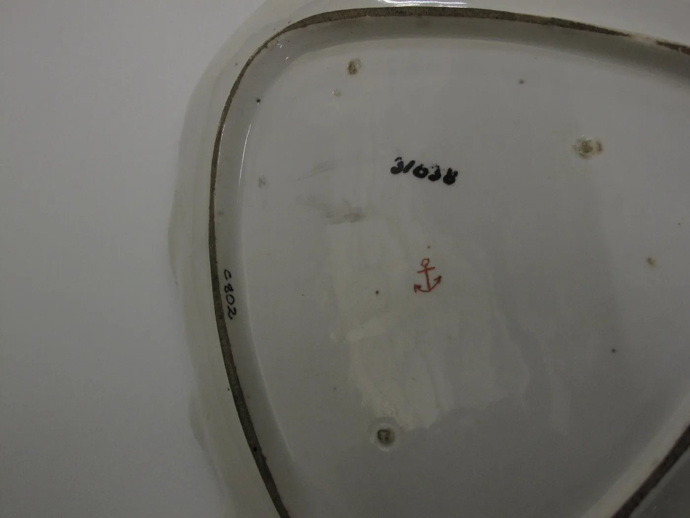 Underside of a white porcelain dish with a small red painted anchor and a black inventory number