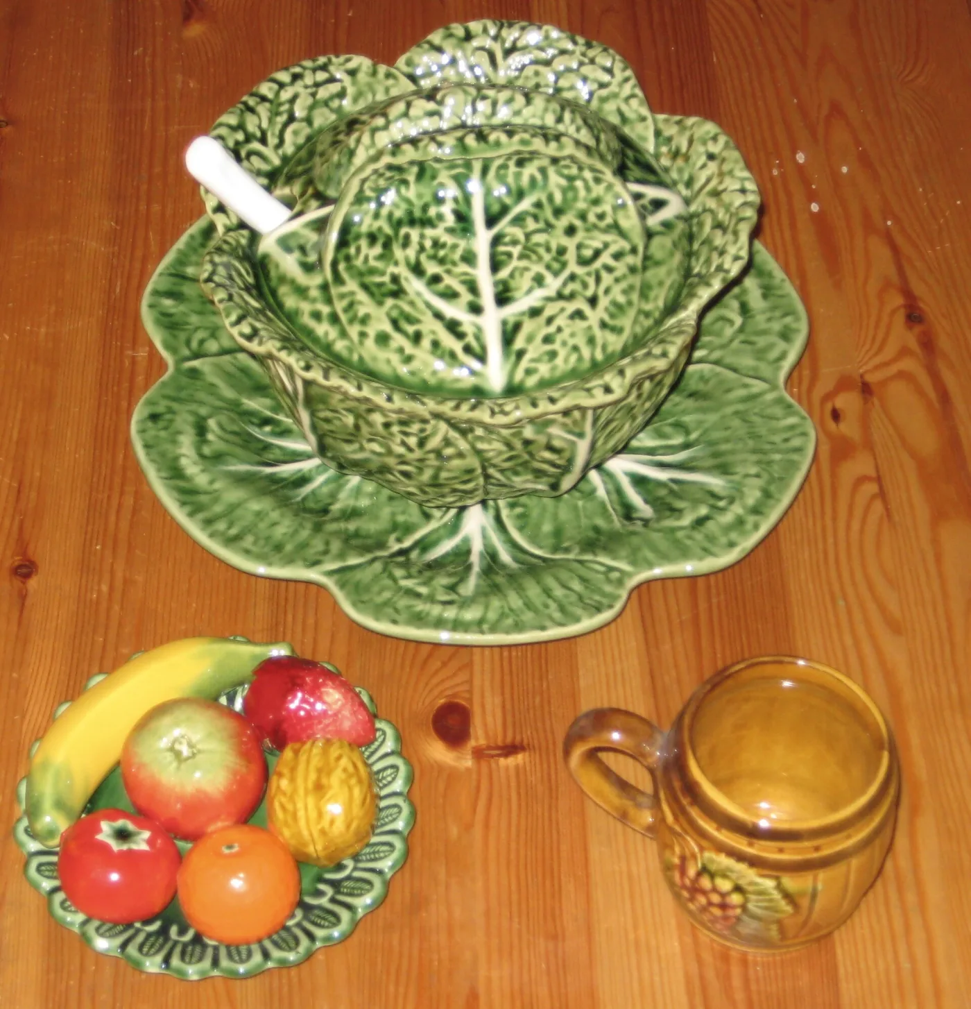 Green cabbage-leaf tureen with matching stand on a pine floor, beside a plate of moulded ceramic fruit and a glazed jug