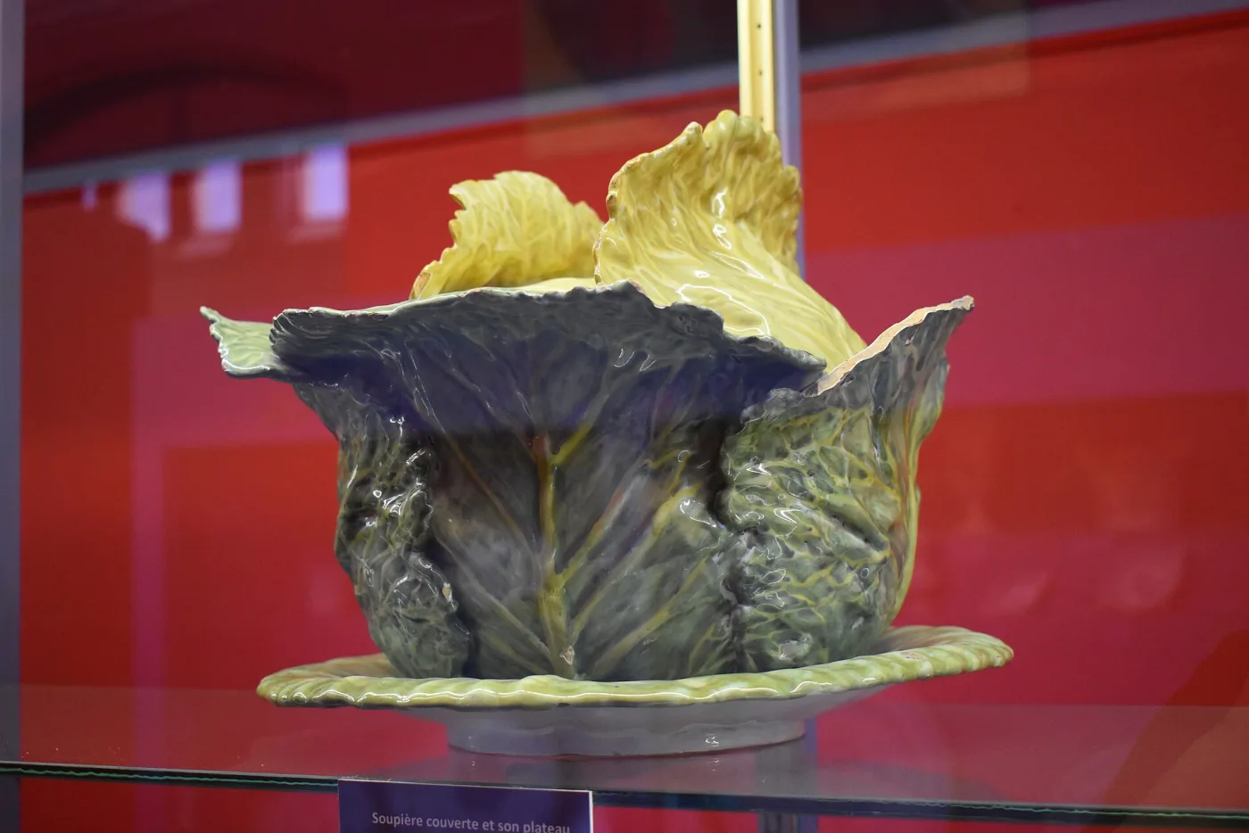 Faience tureen shaped as a headed cabbage with purple-grey outer leaves and yellow inner leaves, on a green leaf stand in a display case