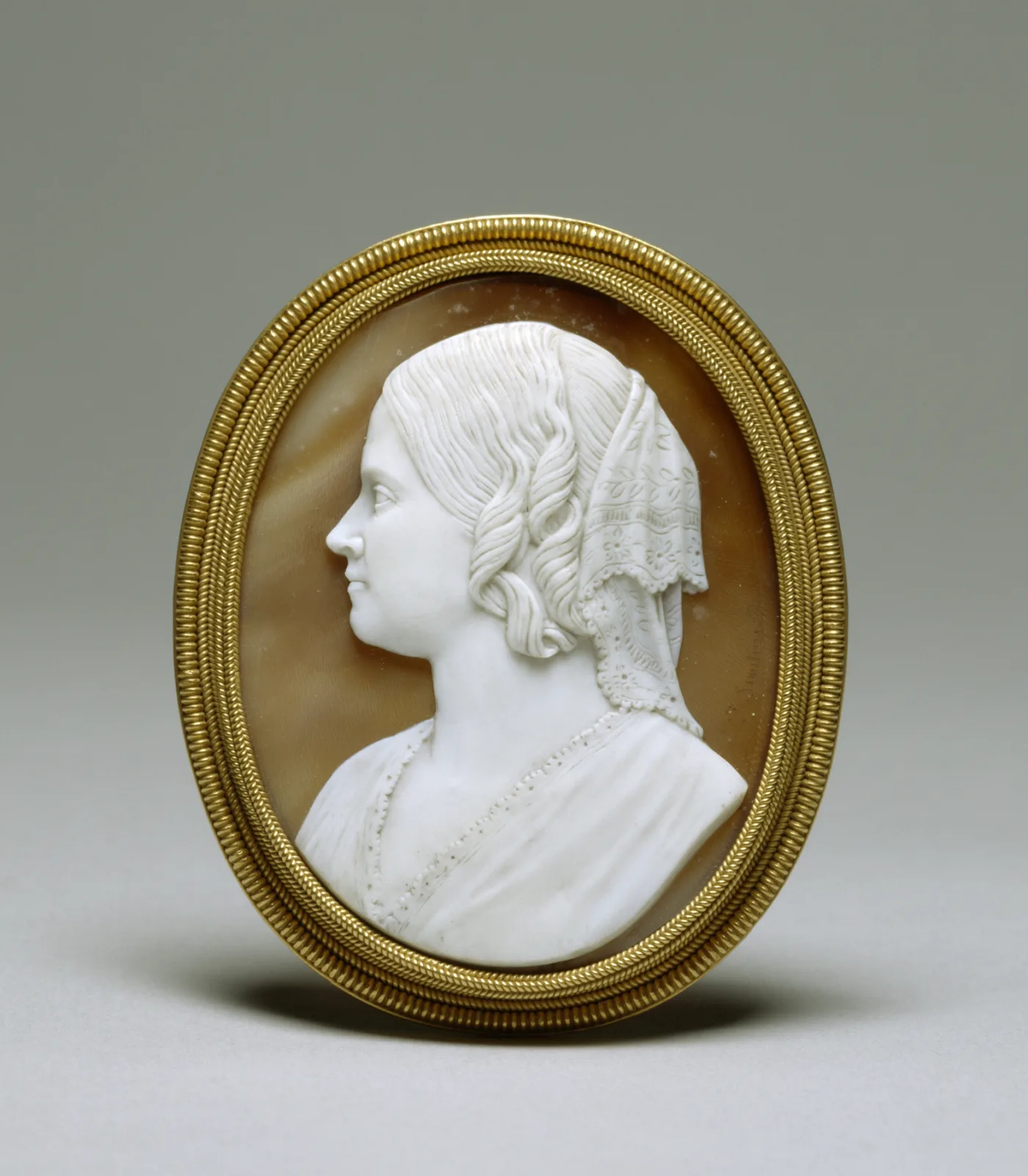 Oval brooch in a plain gold twisted-rope frame, set with a shell cameo of a woman's profile bust in white against a warm caramel-brown ground, wearing a lace mantilla, with a carver's signature incised in the ground at the right