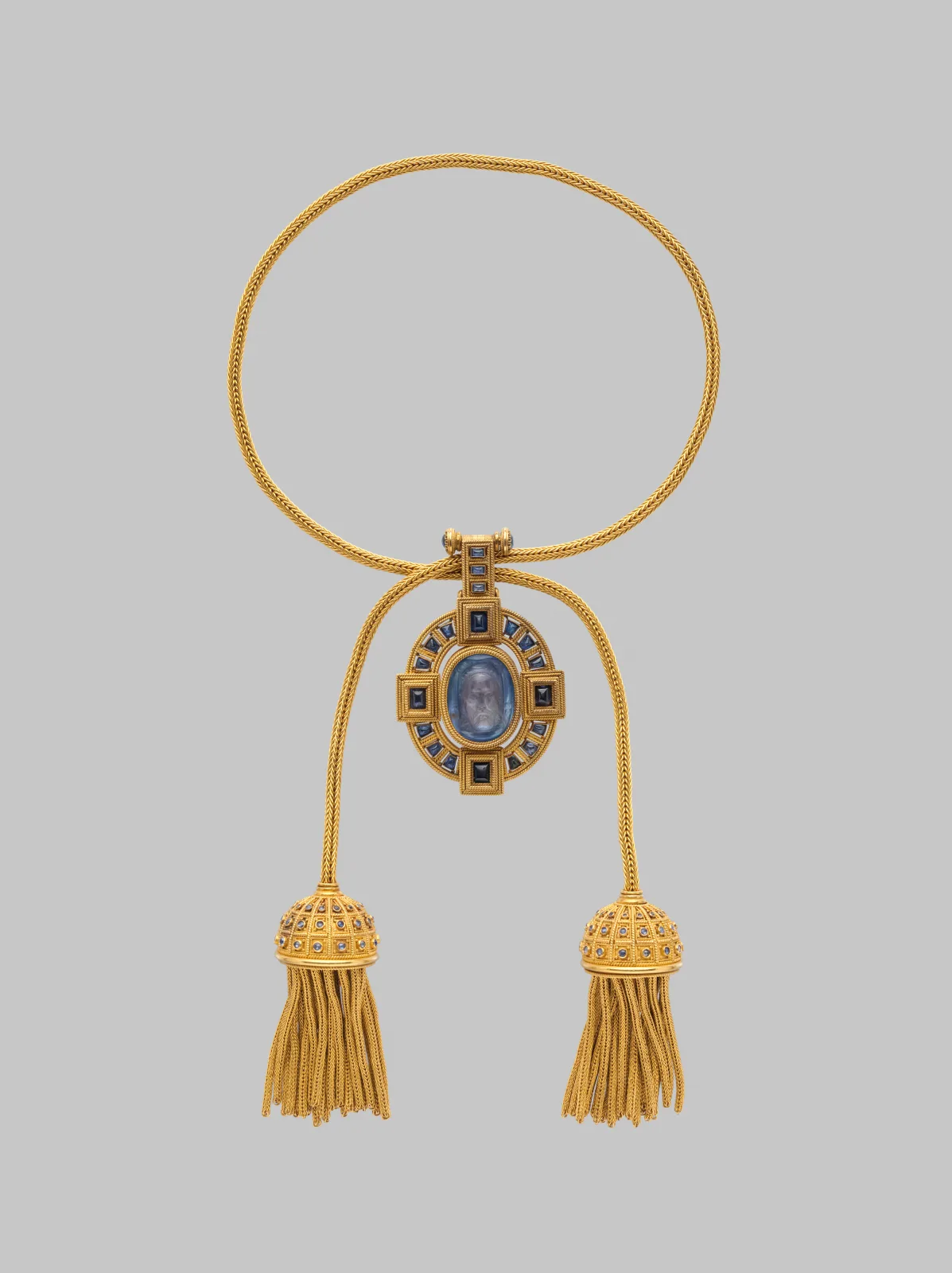 Gold necklace of woven foxtail chain with two tasselled terminals capped in sapphire-set domes, hung with a cruciform pendant of square and rectangular sapphires framing a small oval blue chalcedony cameo of a bearded head