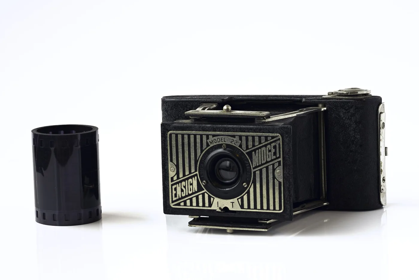 Very small folding camera with a patterned metal faceplate standing beside an upright cylindrical film cassette of comparable height