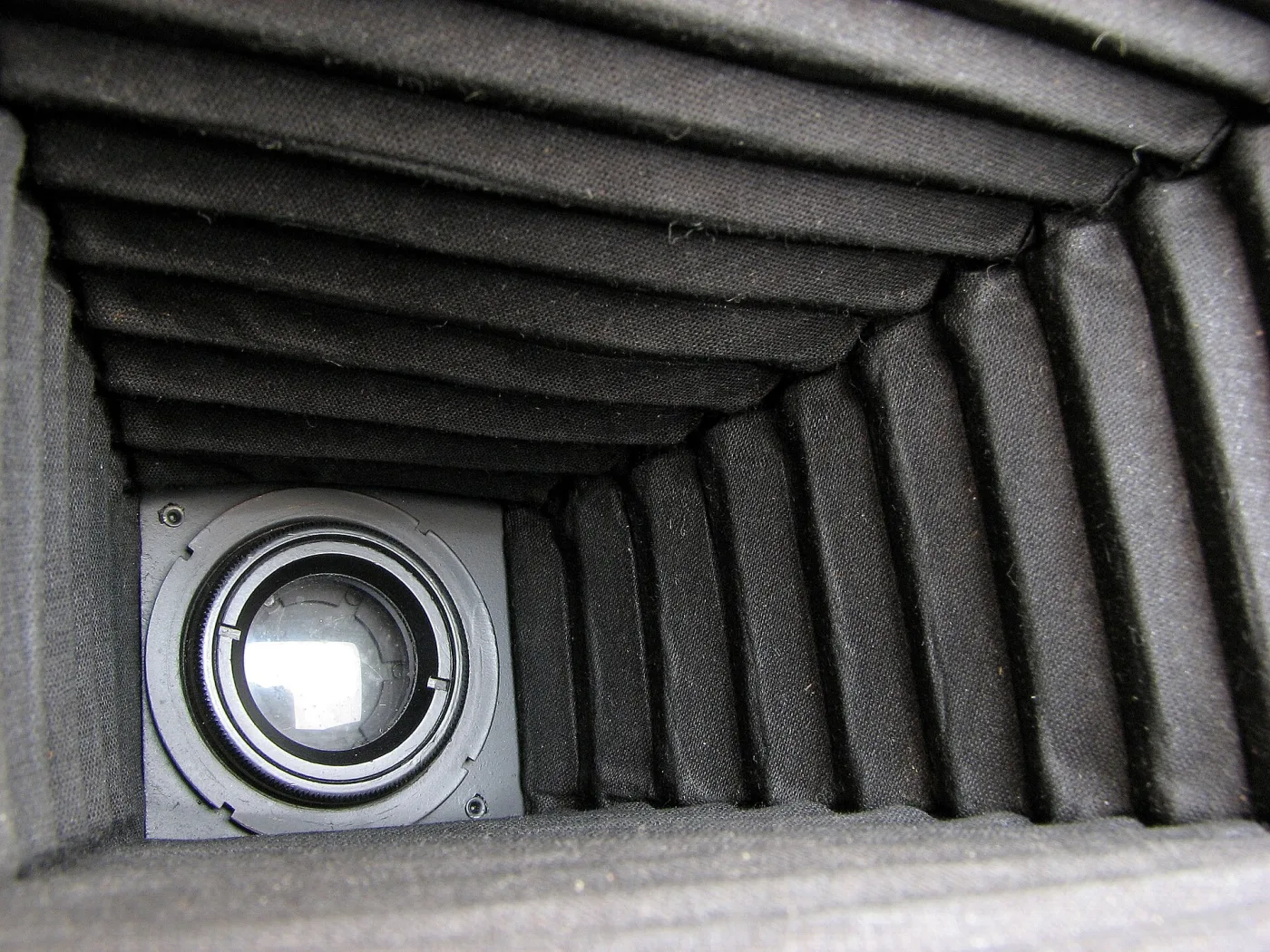 View taken from inside a camera looking down the length of the ribbed black bellows towards the circular lens aperture glowing at the far end