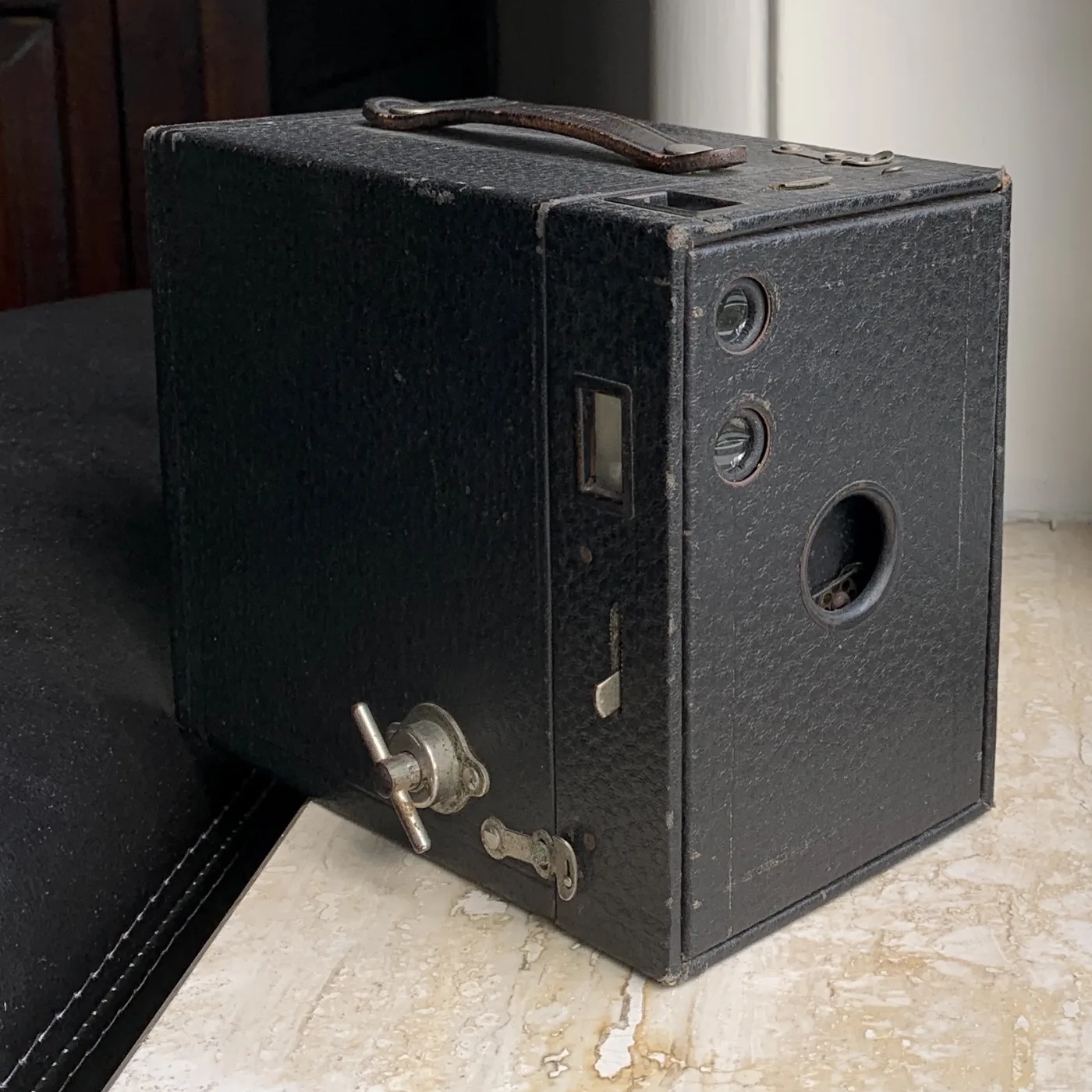 Simple black rectangular box camera of leather-covered card and metal, with a small round lens opening at the front, two brilliant finders and a winding key on the side