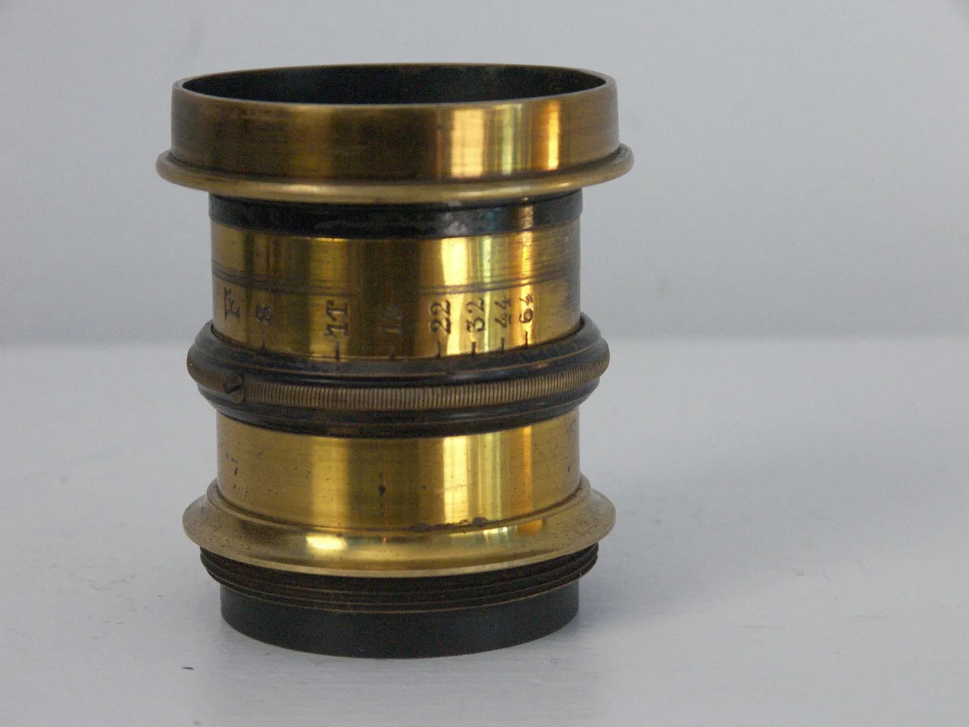 Heavy unmounted brass lens barrel standing on end against a plain background, its polished surface carrying a faint engraved aperture scale and no maker's name