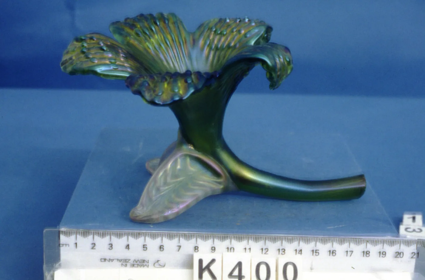 Slender green carnival glass vase with a flared petal-edged rim, lying on its side beside a centimetre ruler