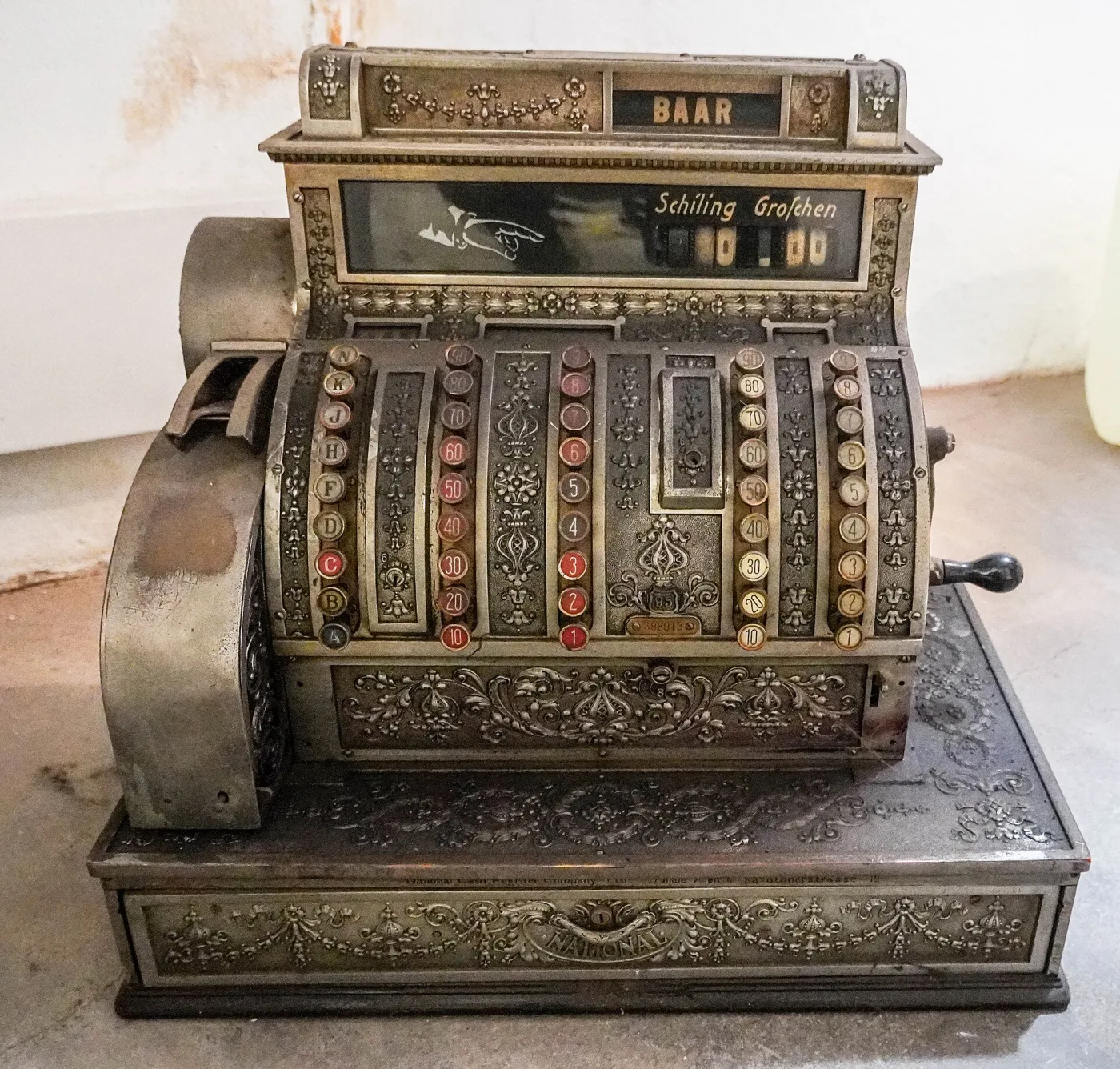 Heavily cast and chased metal cash register with two banks of round keys, a scrolled top sign, and a broad curved drawer front