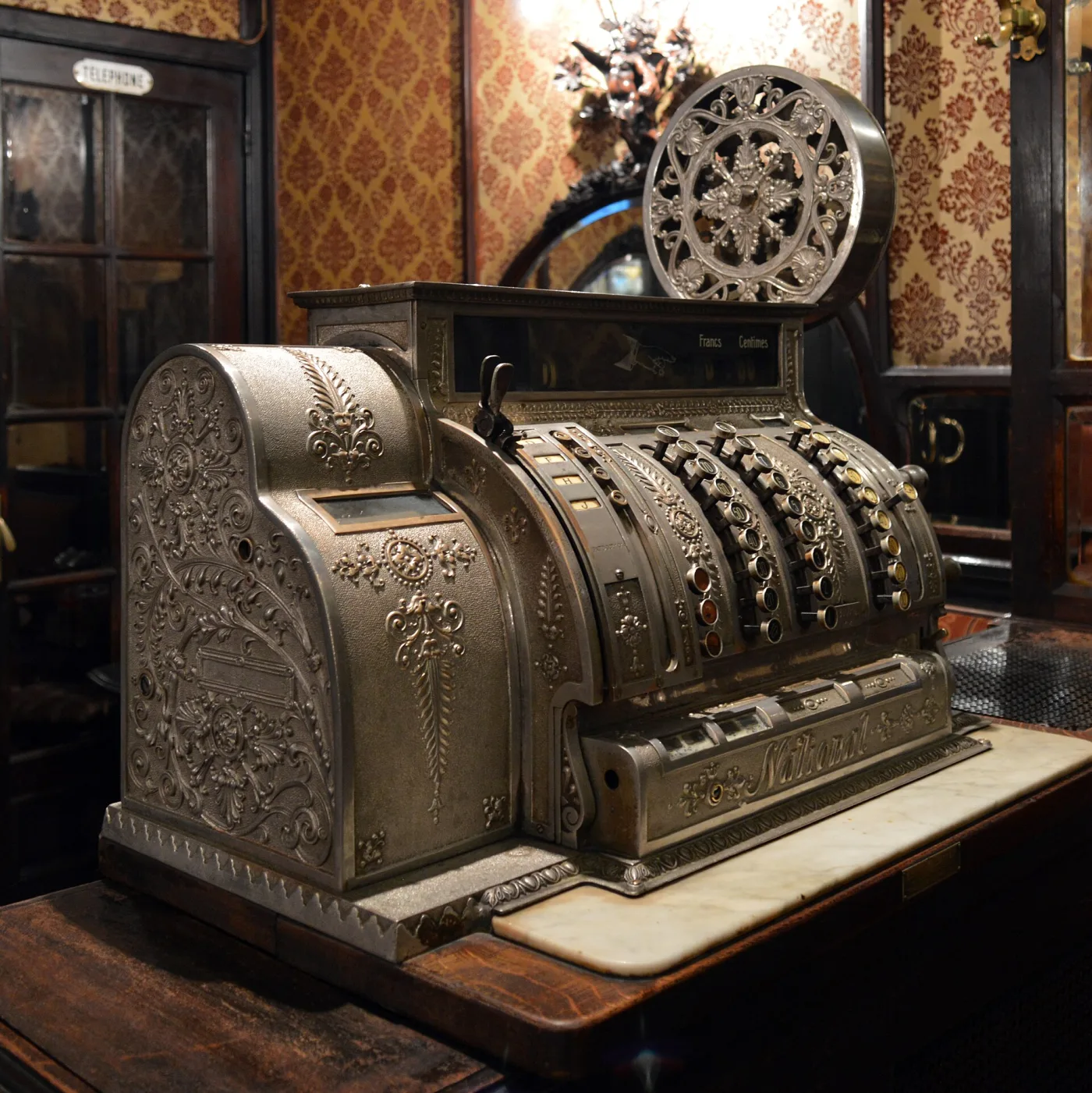 Ornate nickel-plated National cash register standing on a dark wooden bar counter in a period café interior