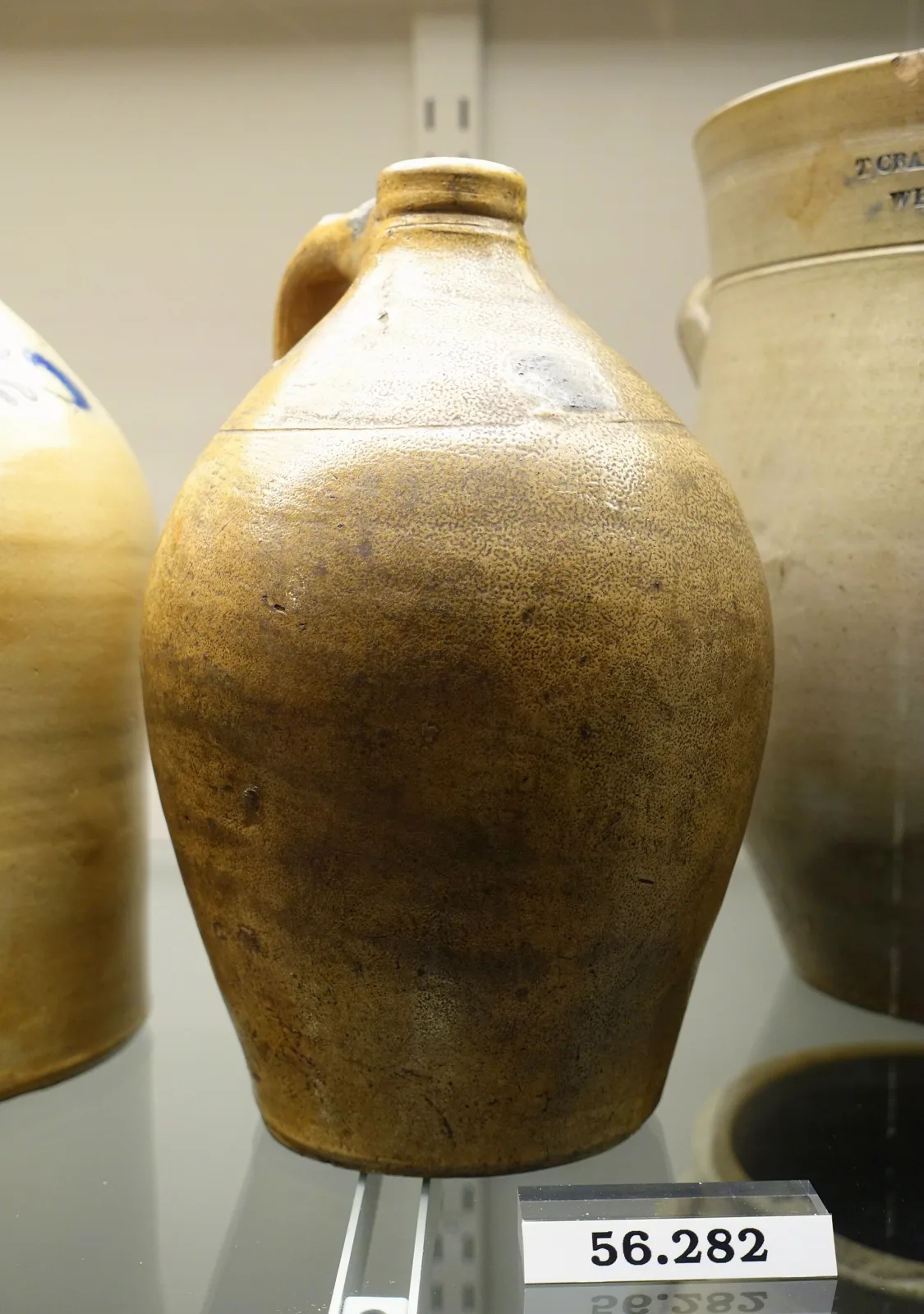 A very similar salt-glazed stoneware jug in the same museum case, its shoulder bare of any mark