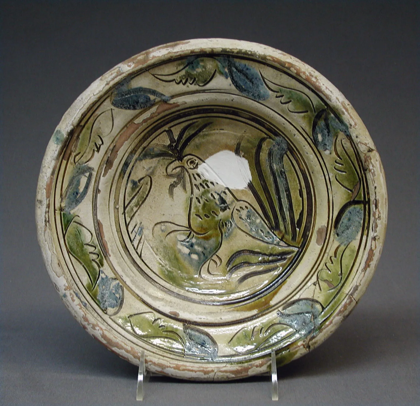 Shallow tin-glazed earthenware dish painted with a green and blue bird at the centre inside a border of stylised leaves, with a large chip missing from the rim