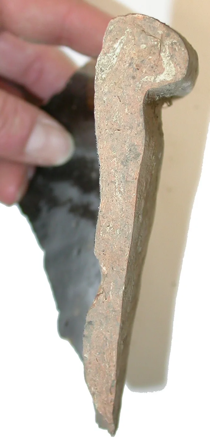 Broken rim fragment of a coarse red earthenware pan held edge-on between finger and thumb, showing the thick clay body and a dark glazed inner surface