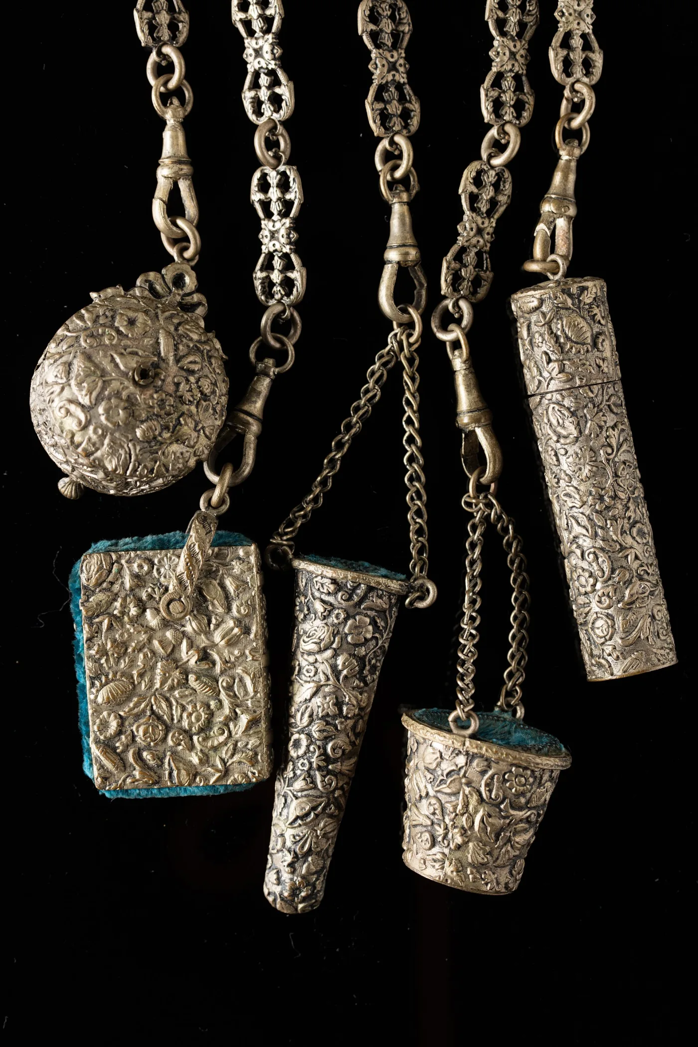 Close view of chatelaine attachments including a circular tape measure with a winding handle, a rectangular pin cushion, a thimble holder and a needle case, all in silver with raised floral decoration
