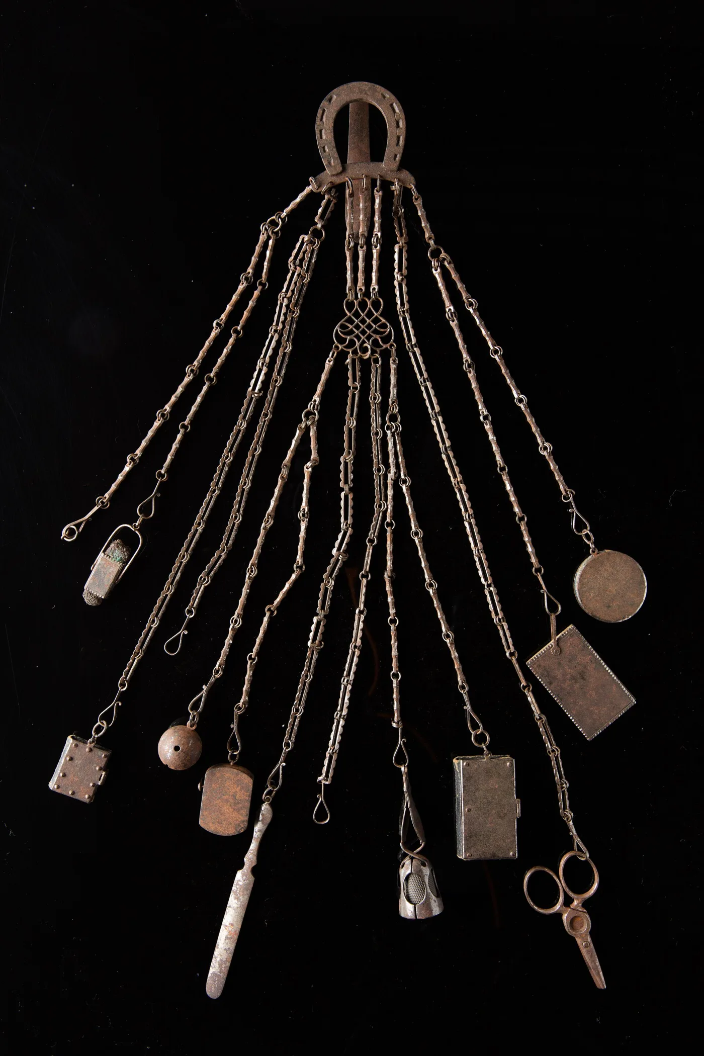 Steel household chatelaine with a horseshoe-shaped belt clip and thirteen chains carrying a notebook, mirror locket, file and thimble