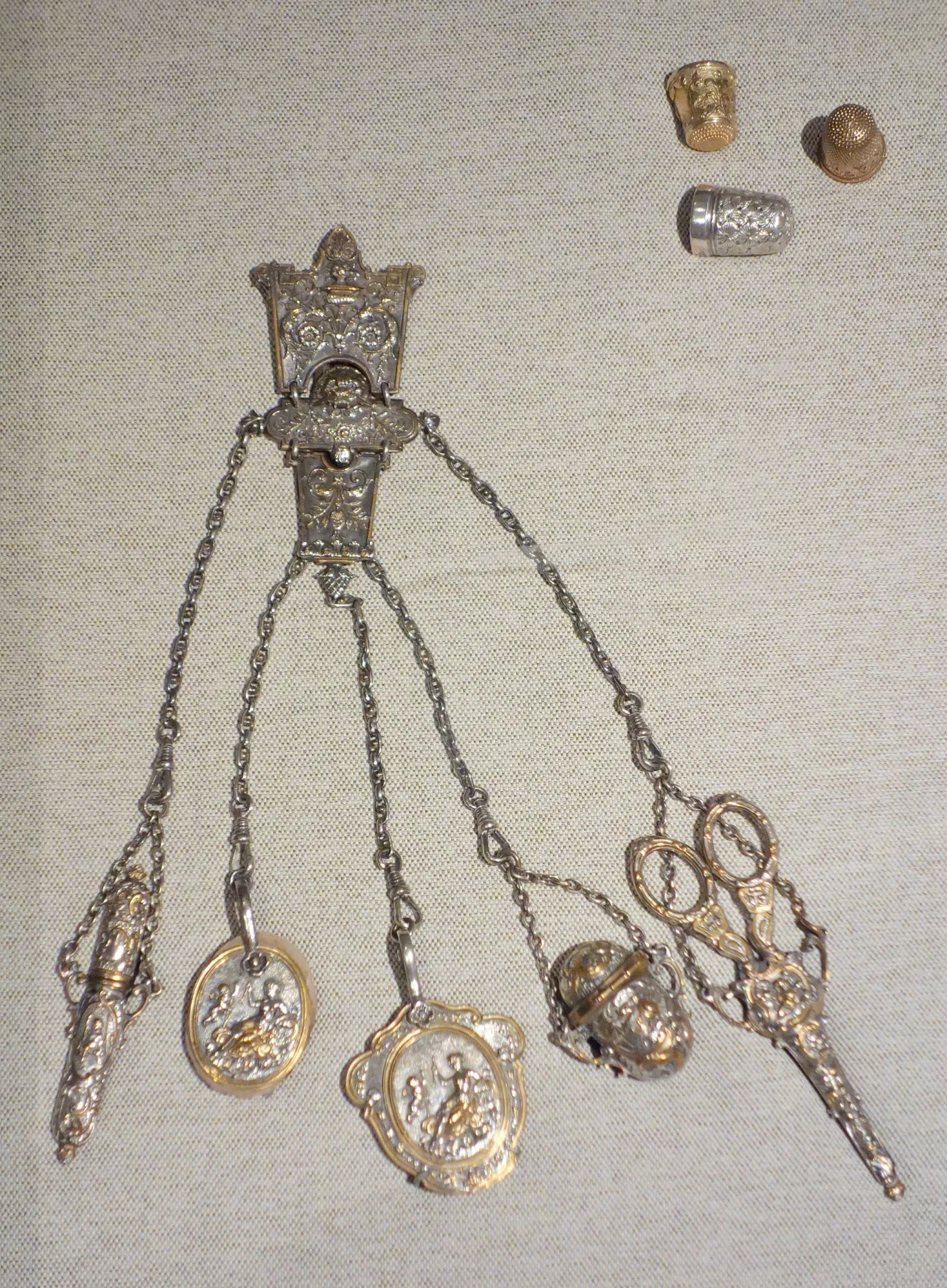 Silver-gilt sewing chatelaine with five chains carrying a needle case, two figural medallions, a thimble holder and gilt scissors, with three thimbles displayed beside it