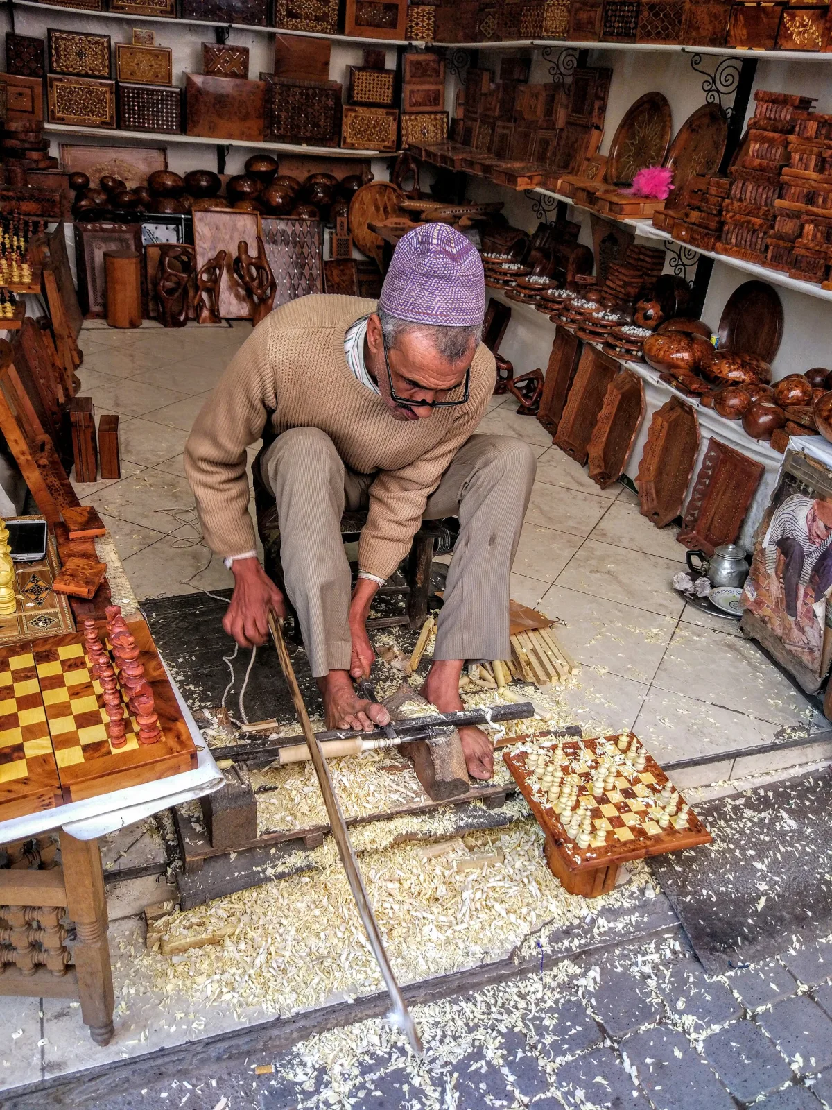 Craftsman crouching over a foot-driven lathe, shaping a chess piece by hand in a workshop lined with finished boards and pieces