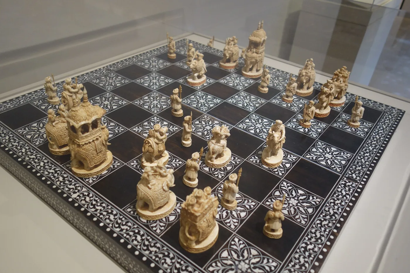 Complete ivory chess set of small carved figures arranged on a dark wooden board inlaid with ivory squares, in a museum case