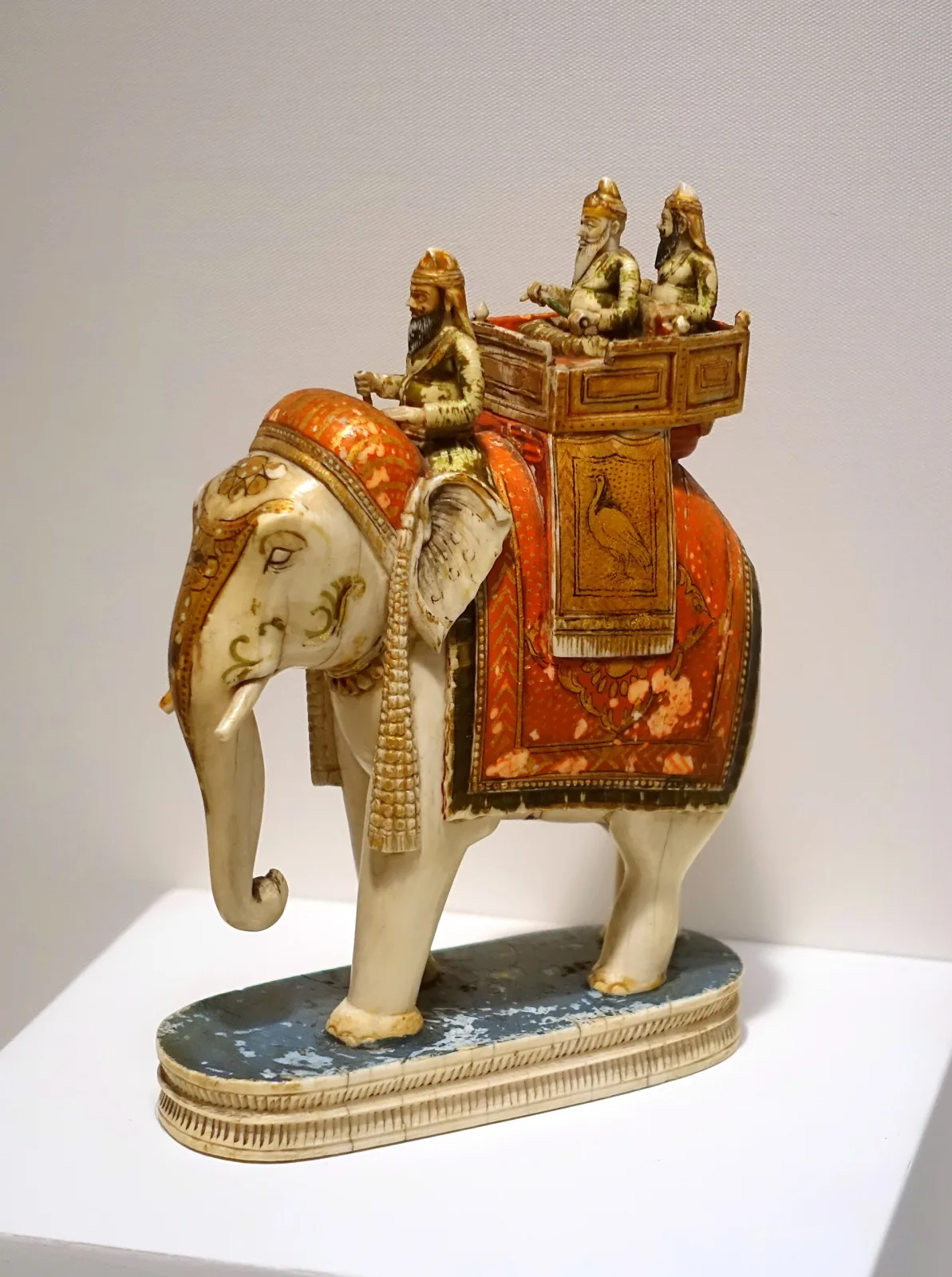 Carved ivory chess piece of an elephant carrying a howdah with riders, painted in red and gold on a blue base