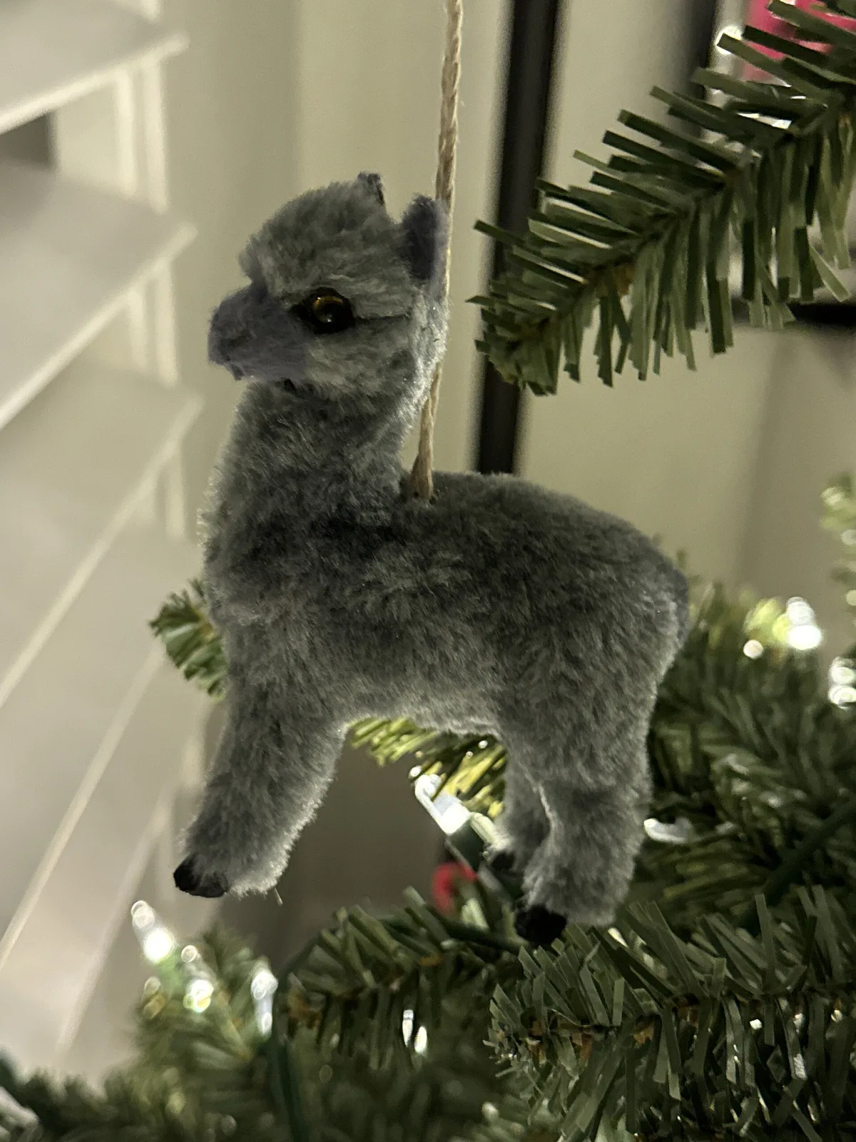 Small standing animal figure made of grey spun cotton over a wire frame, with dark glass eyes and slender legs, hung by a thread from a fir branch