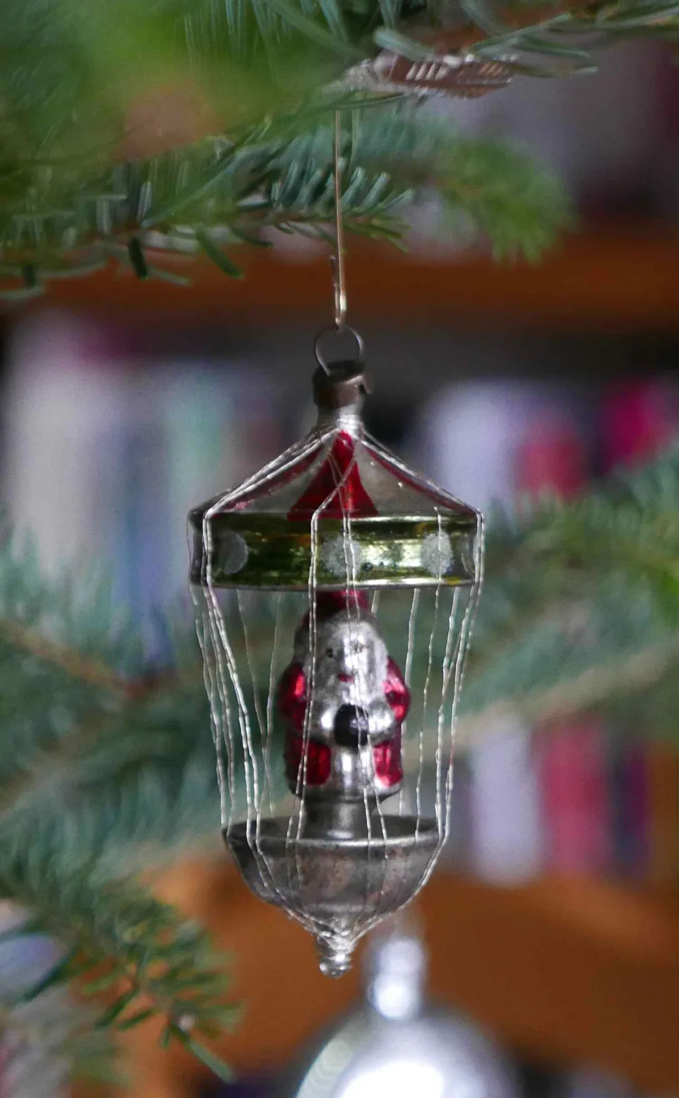 Glass lantern ornament with a small silvered figure suspended inside a cage of fine twisted metal rods, hanging from a fir branch