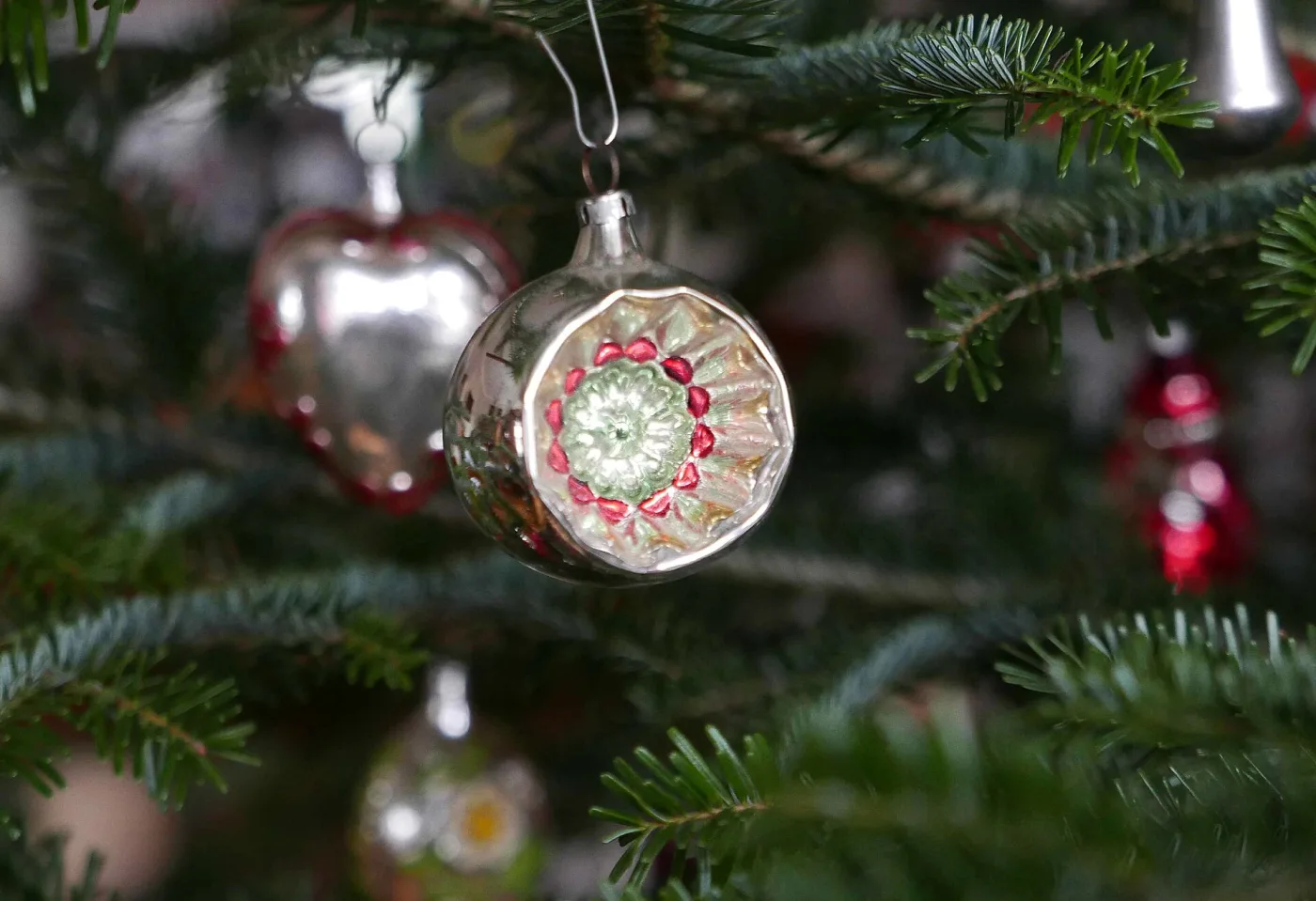 Several silvered indent ornaments hanging close together on a fir tree, their metal caps and wire hangers visible against the branches
