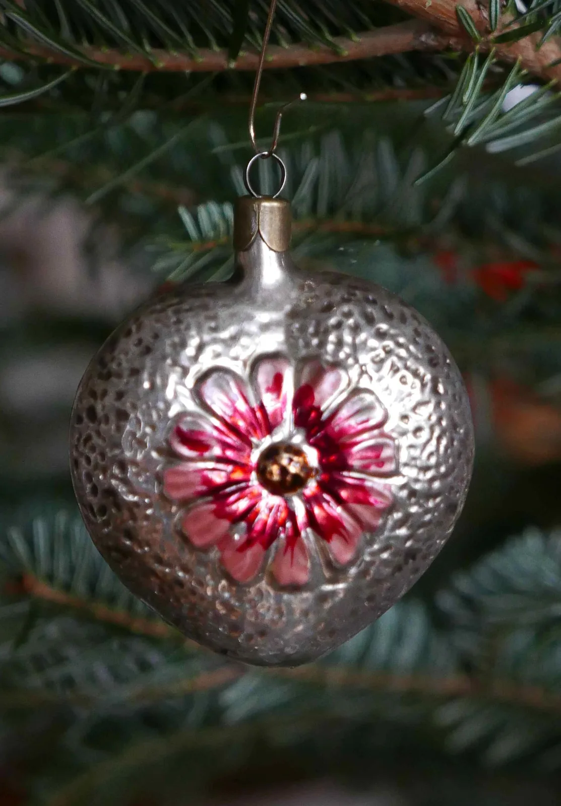 Silvered glass ornament with a finely pebbled surface and a deep round indent holding a red and gold moulded flower, hanging on a fir branch