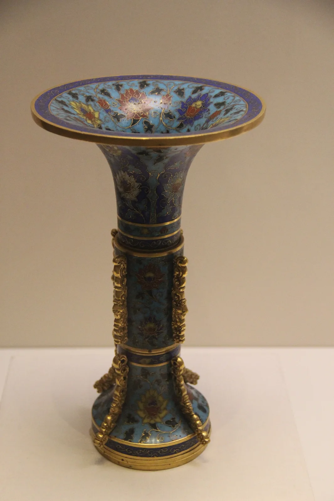 A tall trumpet-mouthed cloisonne vessel with a turquoise ground and coloured floral scrollwork, shown in a museum case