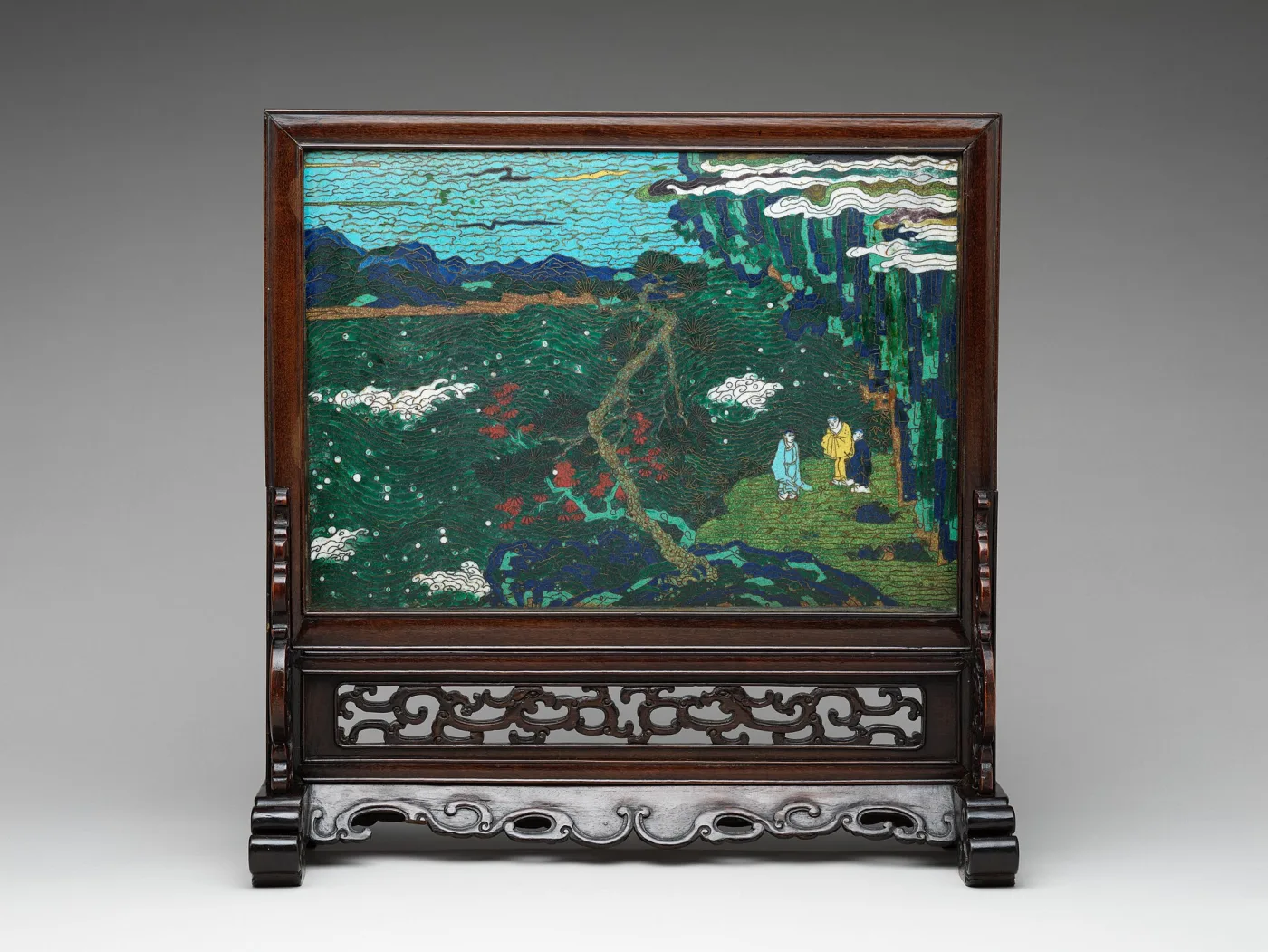A rectangular cloisonne panel showing a landscape with small figures, mounted in a carved wooden frame and stand