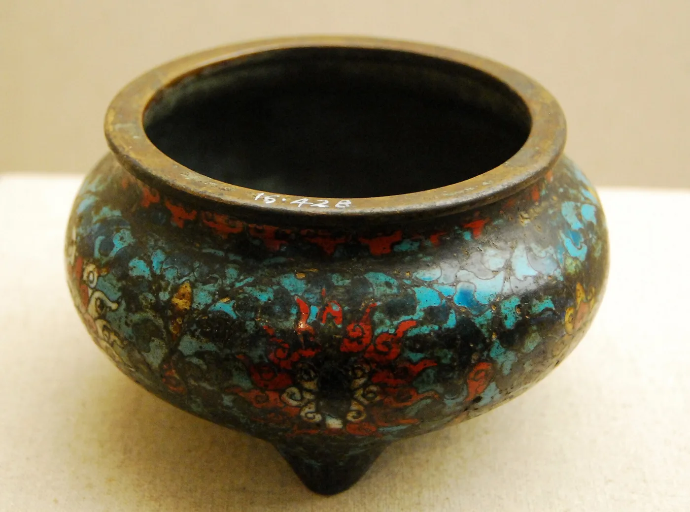 A small three-footed cloisonne censer with worn turquoise, red and white scrolling decoration on a dark ground, an old number painted in white on its rim