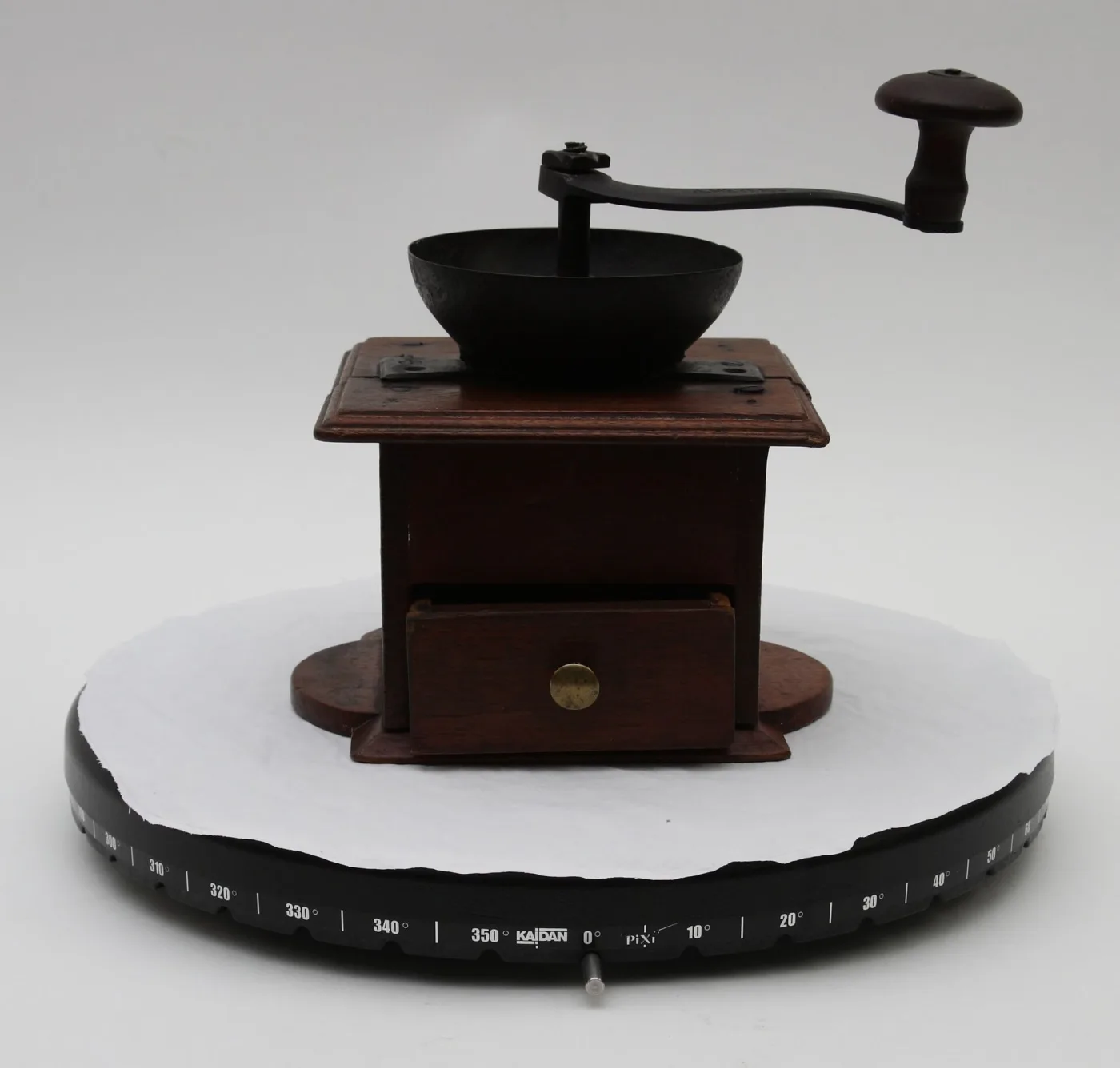 Walnut box coffee mill with an iron funnel hopper and crank on top and a small brass-knobbed drawer at the front, standing on a calibrated photographic turntable