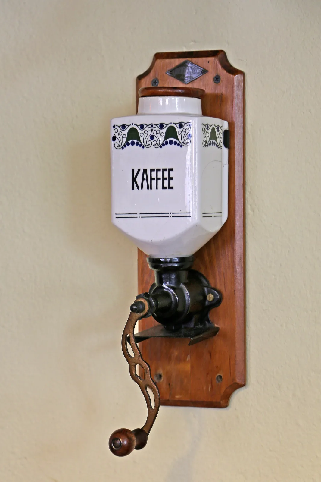 Wall-mounted coffee mill on a shaped wooden backboard, with a white ceramic hopper lettered Kaffee above an iron grinding body and crank