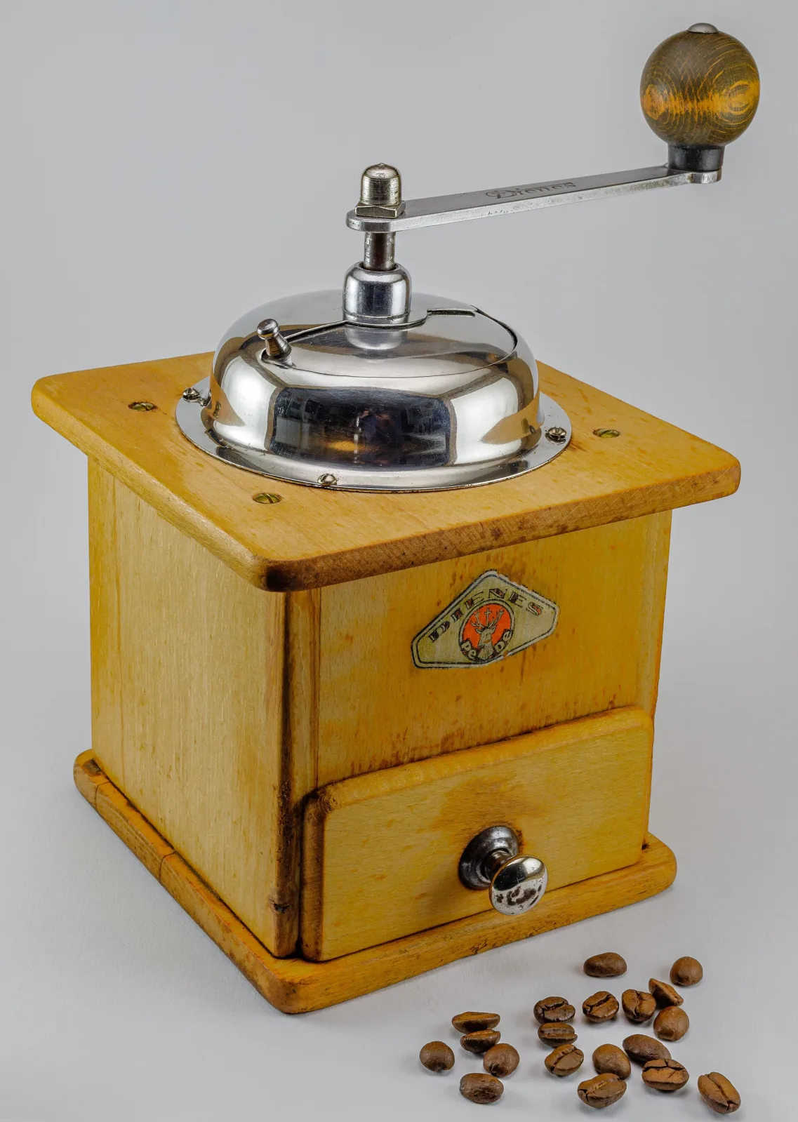 Pale yellow wooden box coffee mill with a domed chromed cap and a long steel crank, a small enamel maker's badge on the front above the drawer, with roasted beans scattered at its foot