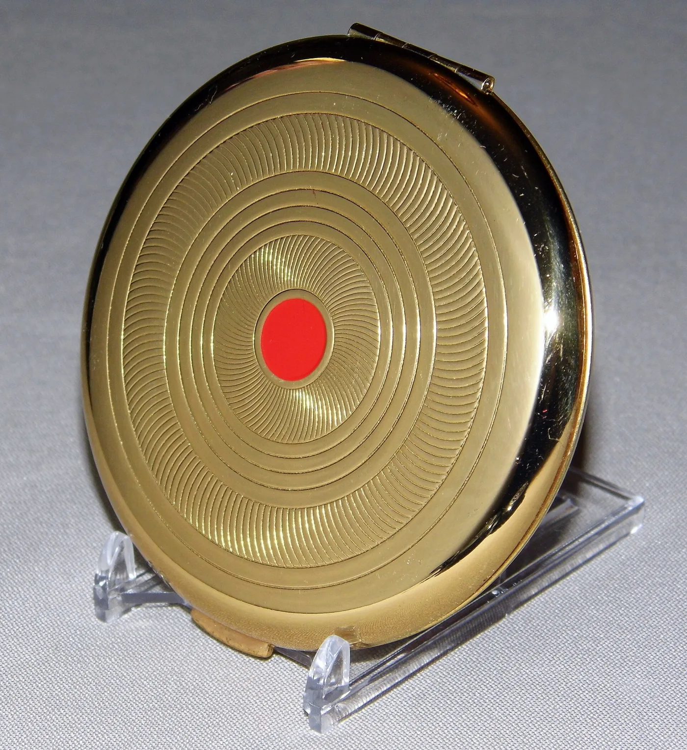 Round gold-tone compact seen from above on a small stand, its lid turned with fine concentric rings around a single flat red enamel disc at the centre, the hinge visible at the back