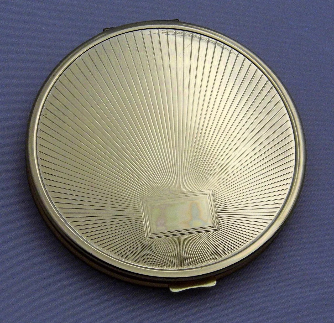 Round pale silver-coloured compact whose lid is engine-turned with fine straight lines radiating from an off-centre point, with a small blank rectangular cartouche near the lower edge