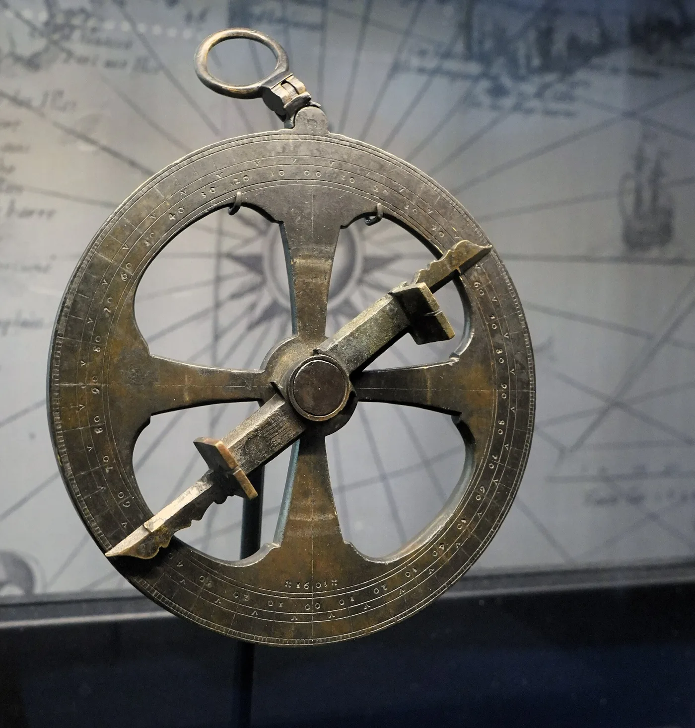 Heavy dark bronze disc pierced by four large openings around a solid centre, carrying a pivoted sighting bar with a raised vane at each end and a suspension ring at the top, mounted against a map