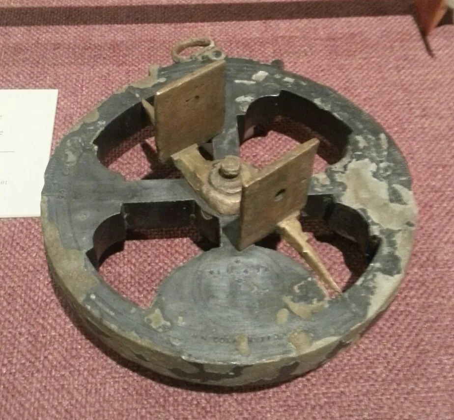 Heavily corroded grey metal astrolabe wheel with four openings, its surface pitted and encrusted, carrying a brighter brass sighting bar with two pierced vanes and a suspension ring at the top