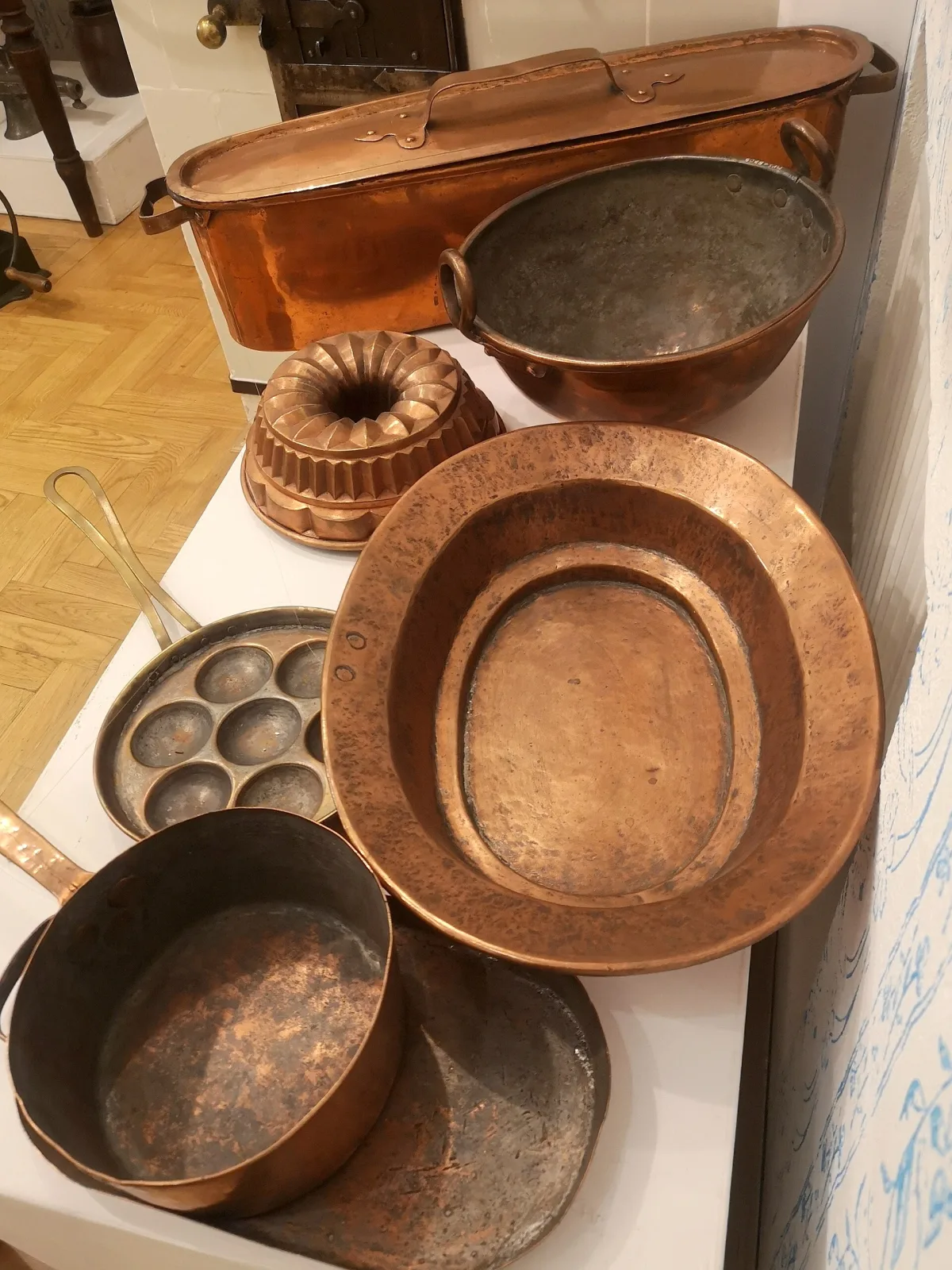 Group of copper vessels on a white plinth: a long lidded fish kettle, a deep bowl with riveted ring handles and a dull grey lining, a fluted ring mould, a wide hammer-marked oval dish with a stepped well, a pan with seven round hollows and a plain deep saucepan with a long handle