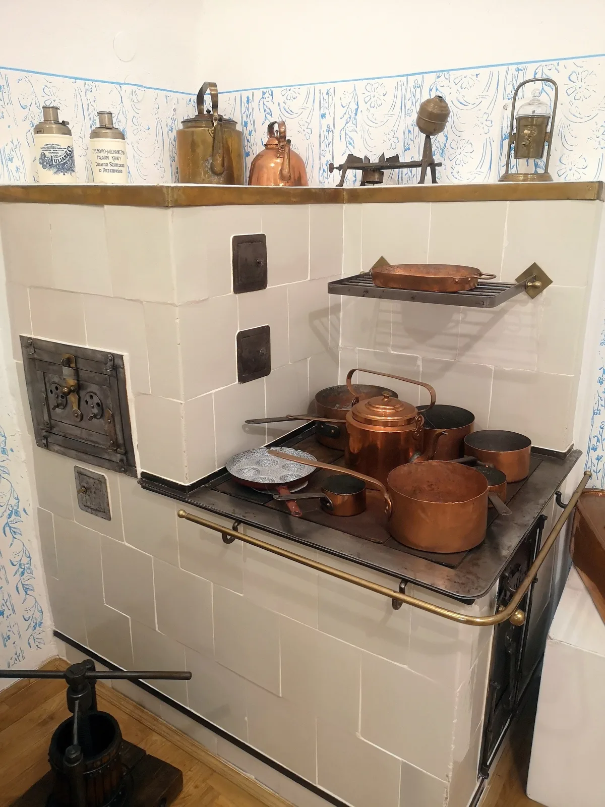 Corner of an old tiled kitchen with a black cast-iron range, on and around which stand several copper pots and pans, their surfaces dull brown rather than polished, with jars and utensils on a shelf above