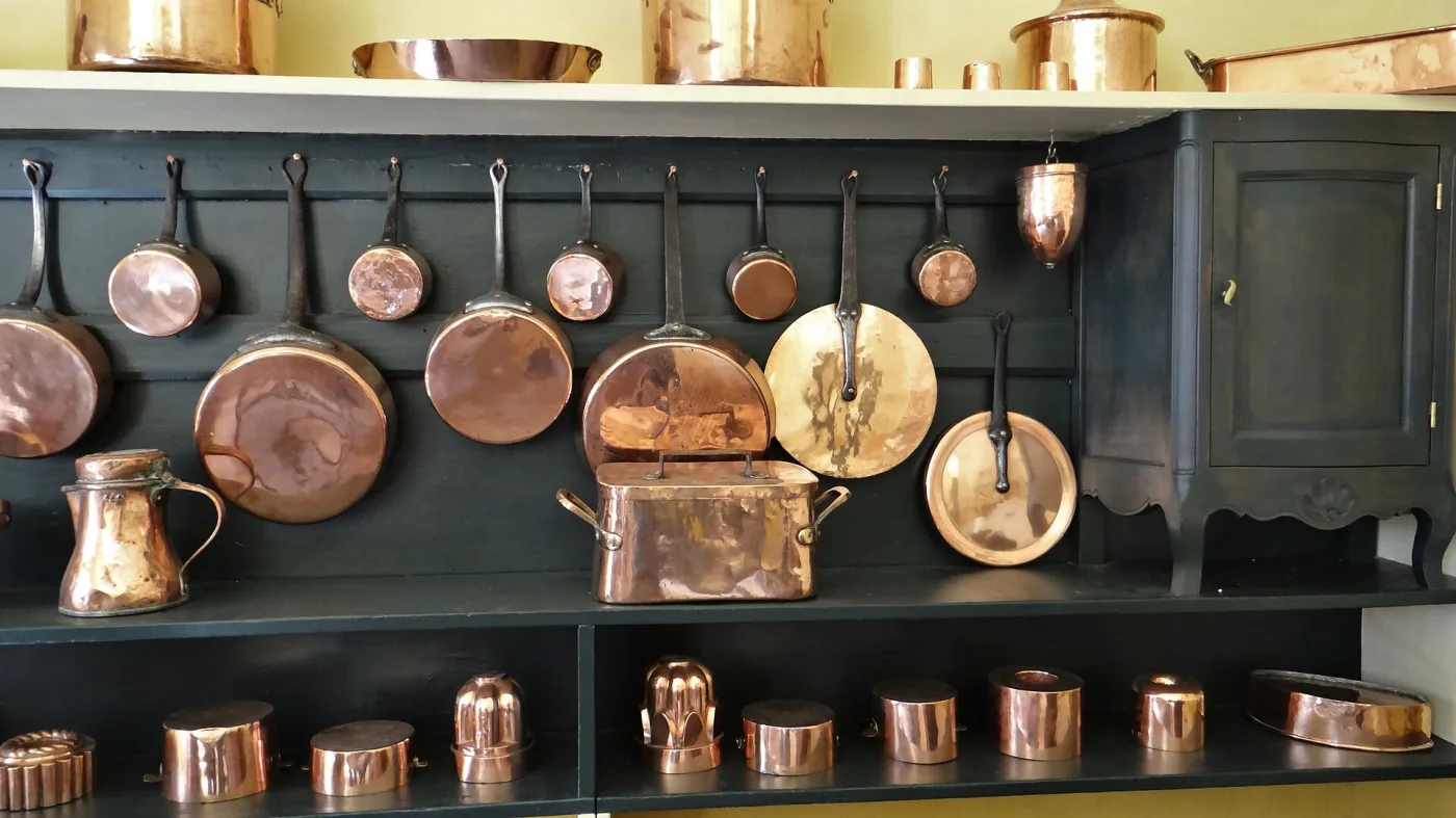 Long dark green kitchen dresser hung with a rail of copper pans of graduated sizes on iron handles, a rectangular two-handled lidded copper braising pan and a copper jug on the shelf, and a row of small copper pots and moulds below; one hanging disc is distinctly paler and yellower than the rest