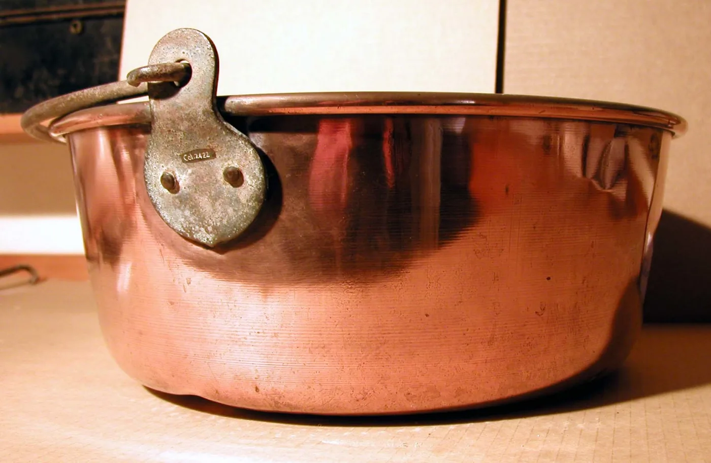 Wide shallow copper preserving pan photographed close, with a heavy iron swing handle hanging from a shaped brass lug fixed to the rim by two large rivets