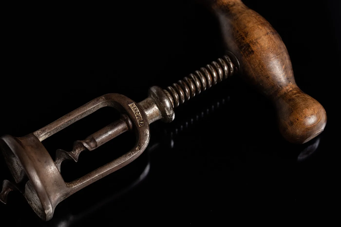 Steel corkscrew with a turned wooden crossbar handle on a threaded shaft running down through an open two-bar frame that ends in a flanged collar, lit against black